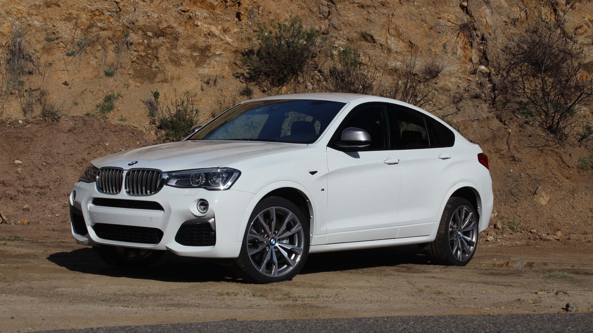 Bmw X4 桌布 壁紙 — 一架時尚的白色BMW X4轎跑SUV停泊在塵土飛揚的路邊，背景是崎嶇的棕色懸崖和稀疏的綠色灌木。
