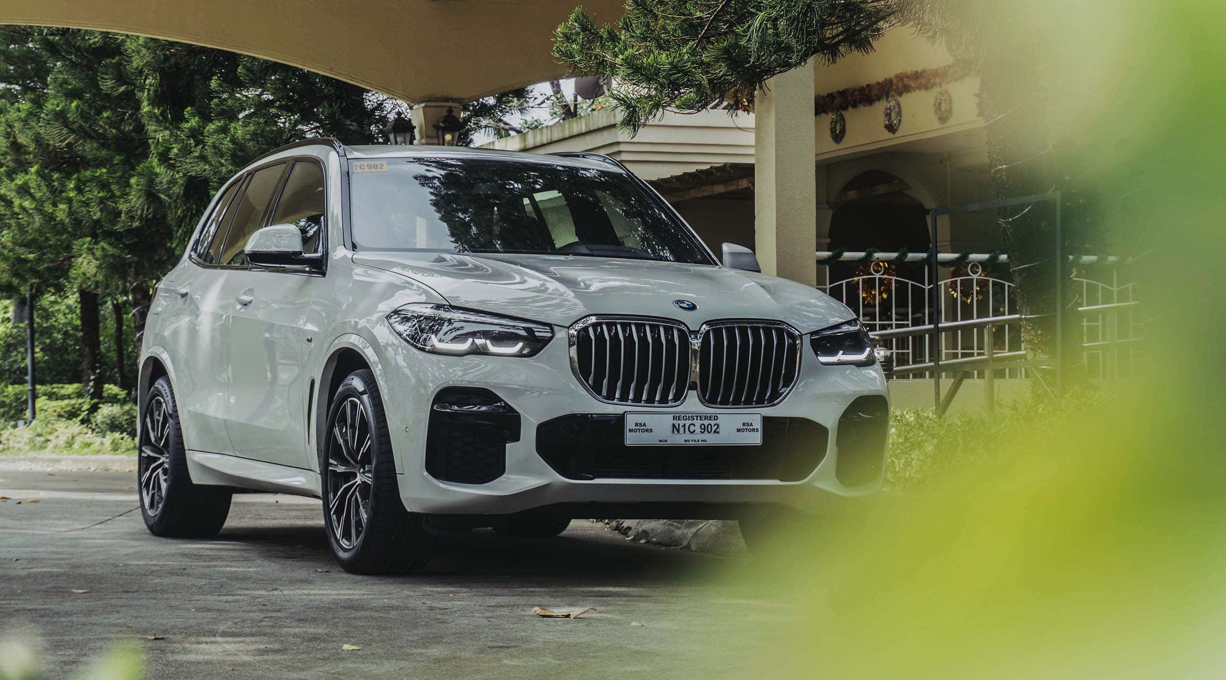 Bmw X5 桌布 壁紙 — 一架時尚的白色BMW X5 SUV停泊在有蓋車道下，周圍環繞著茂密的綠植和一棟經典房屋。