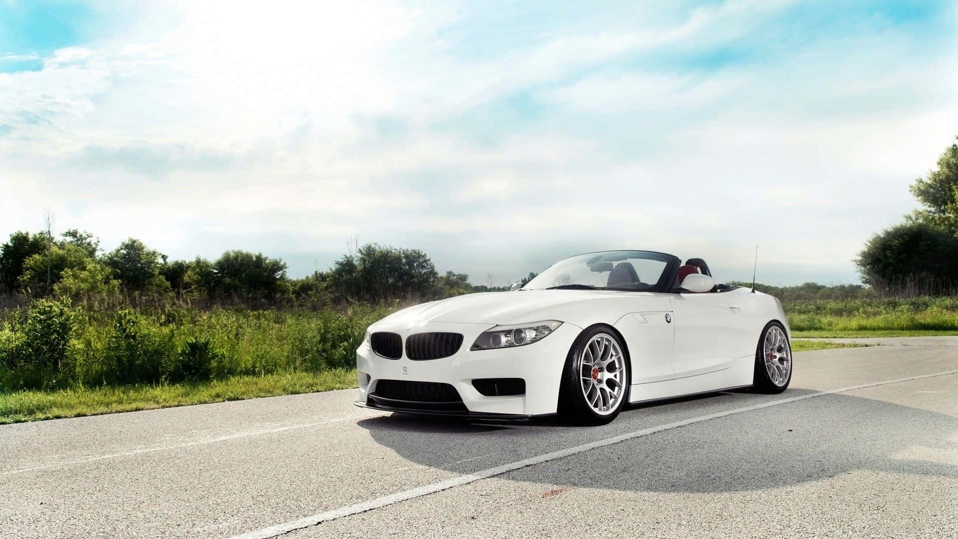Bmw Z4 桌布 壁紙 — 一架時尚的白色BMW Z4開篷車停泊在陽光明媚的鄉村道路上，周圍有綠色樹葉和部分多雲的天空。