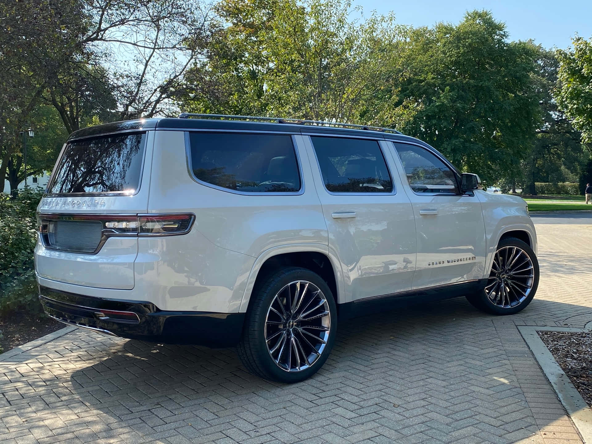 Jeep Wagoneer 桌布 壁紙 — 一架時尚的白色GMC Yukon Denali SUV停泊在陽光普照的磚砌車道上，周圍環繞著茂密的綠樹。