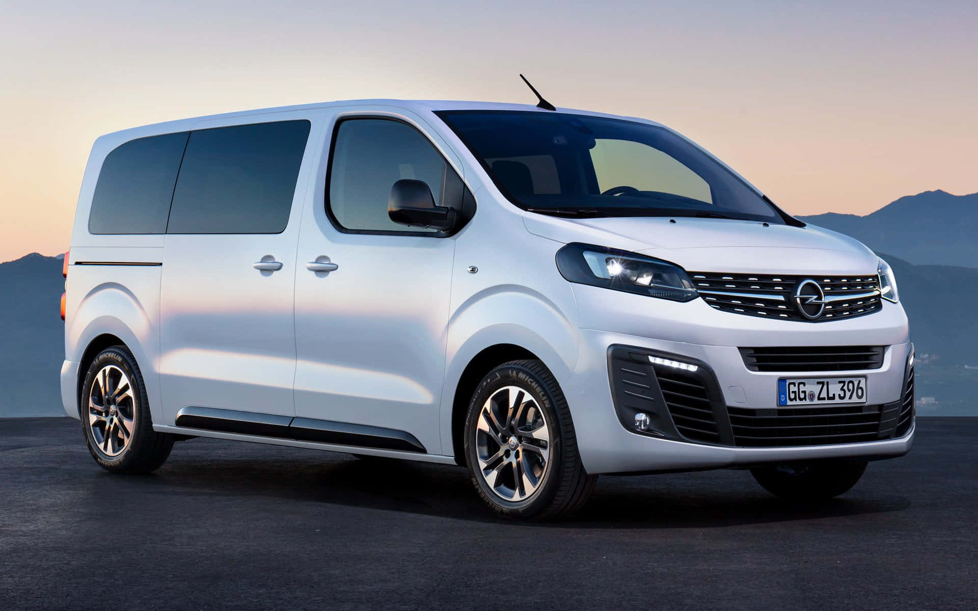 Opel Zafira 桌布 壁紙 — 一架時尚的白色Opel Vivaro客貨車停在深色地面上，背景有山脈和柔和嘅日落天空。