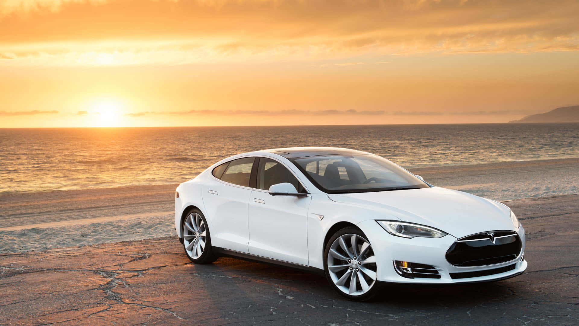 tesla 桌布 壁紙 — 一架時尚的白色Tesla Model S停泊在龜裂的瀝青海灘上，金色夕陽與海浪翻滾。