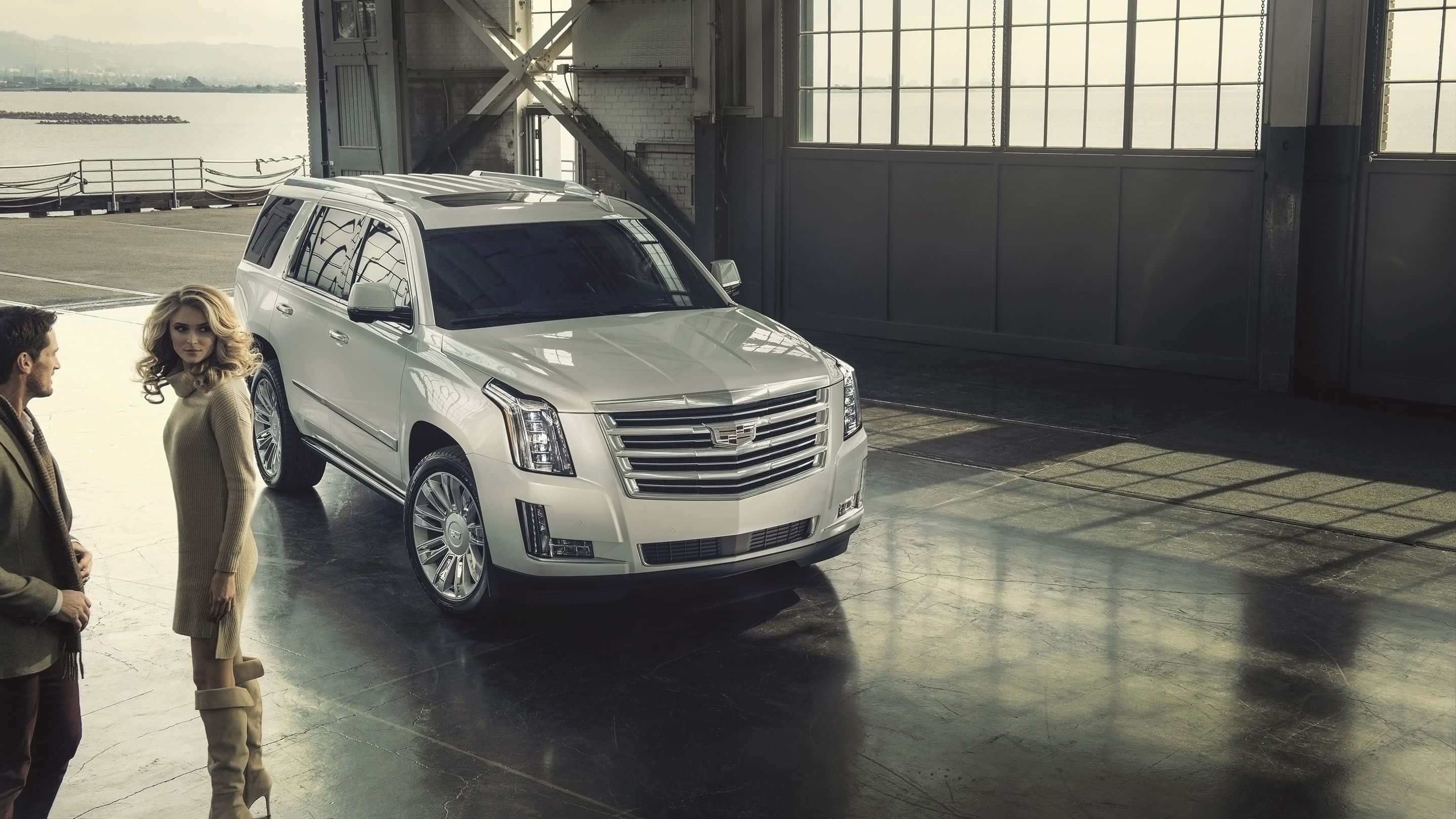Cadillac Escalade 桌布 壁紙 — 一架時尚的白色凱迪拉克SUV在自然光下，於寬敞的工業機庫中閃閃發光，旁邊站著兩位衣著優雅的人。