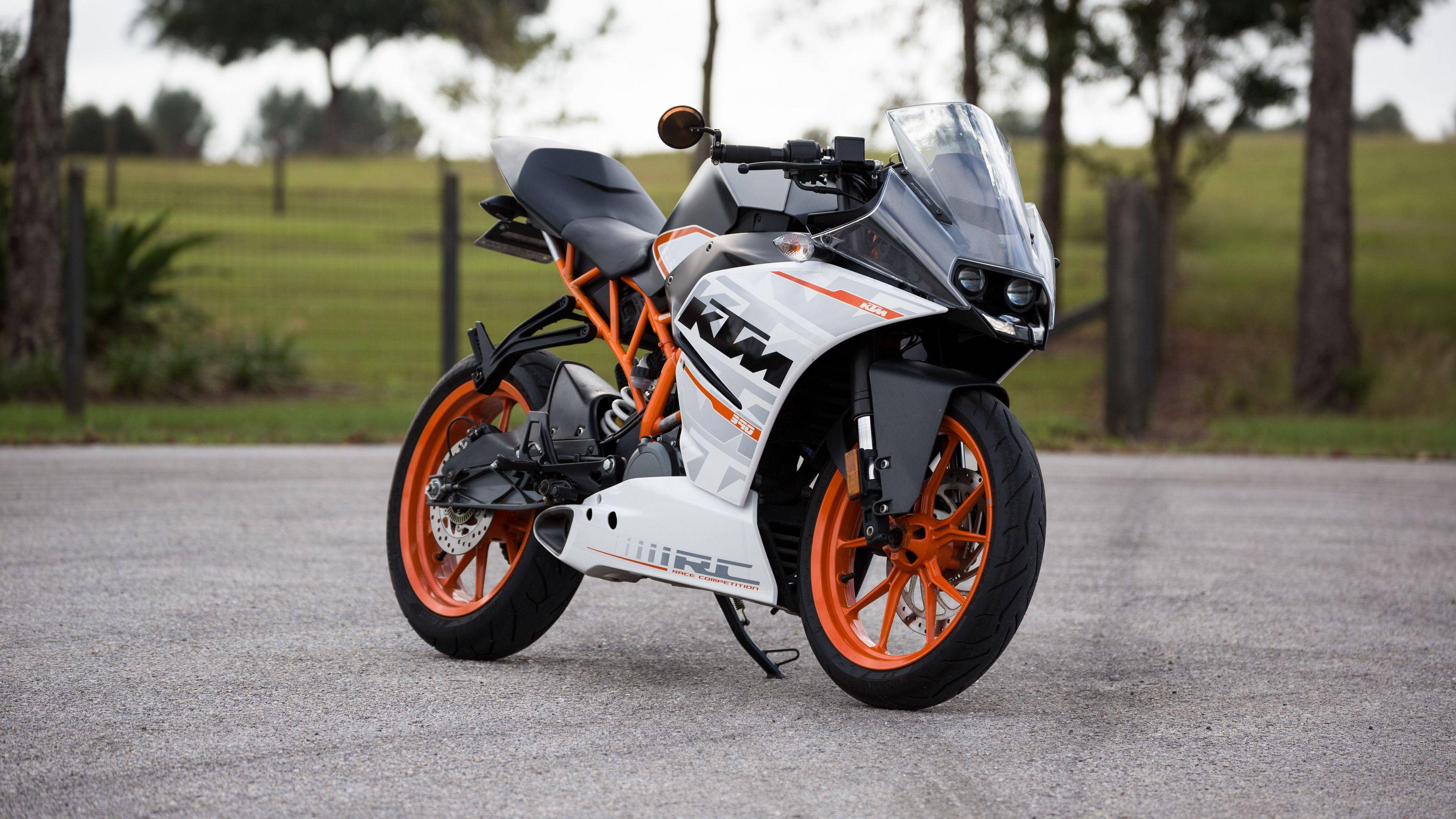 機車 桌布 壁紙 — 一架時尚的白色和橙色KTM RC電單車停在碎石路上，周圍是綠草和樹木。