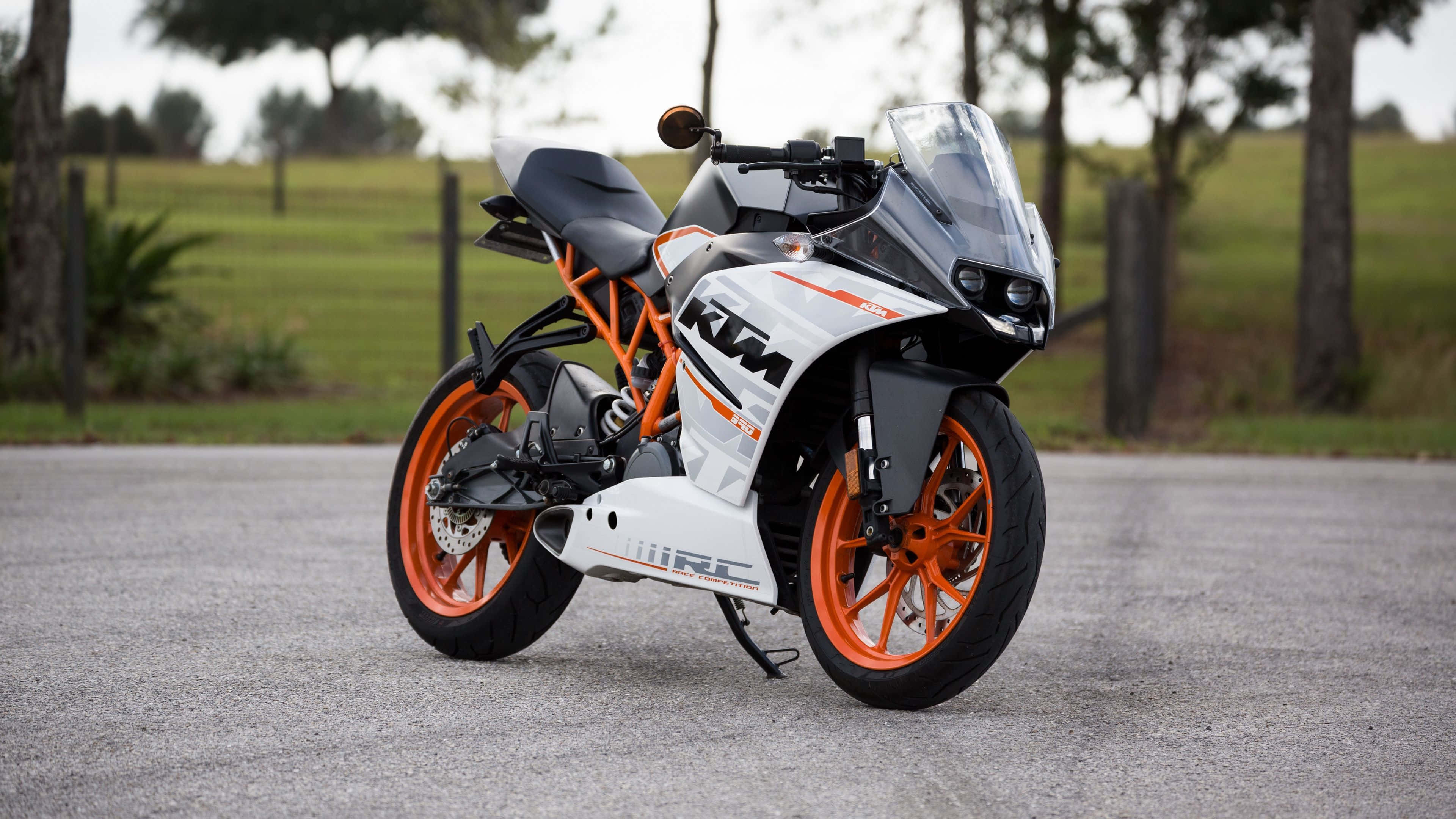 Ktm Bike 桌布 壁紙 — 一架時尚的白色和橙色KTM RC電單車停在碎石路上，周圍是綠草和樹木。