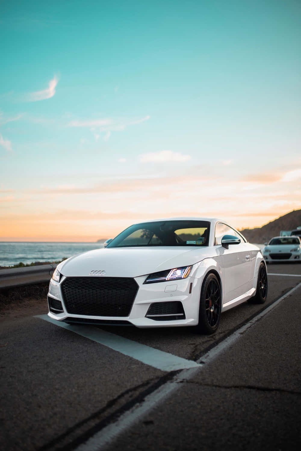 Audi Tt Rs 桌布 壁紙 — 一架時尚的白色奧迪TT轎跑車停泊在日落時分的海岸公路上，背景是海洋和山丘。