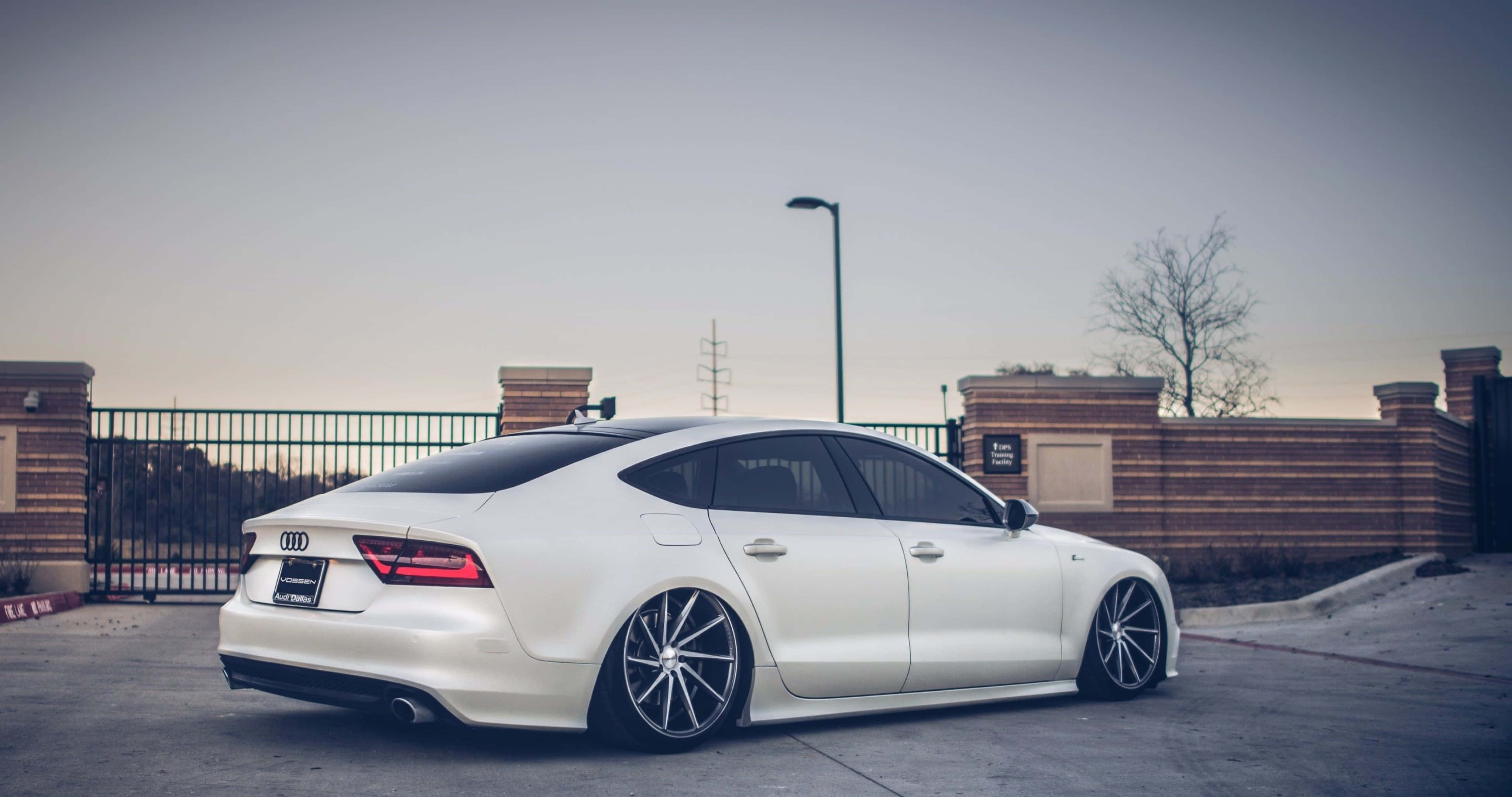 Audi S7 桌布 壁紙 — 一架時尚的白色奧迪房車配備訂製輪圈，停泊在黃昏時分的混凝土停車場，靠近磚牆和圍欄。