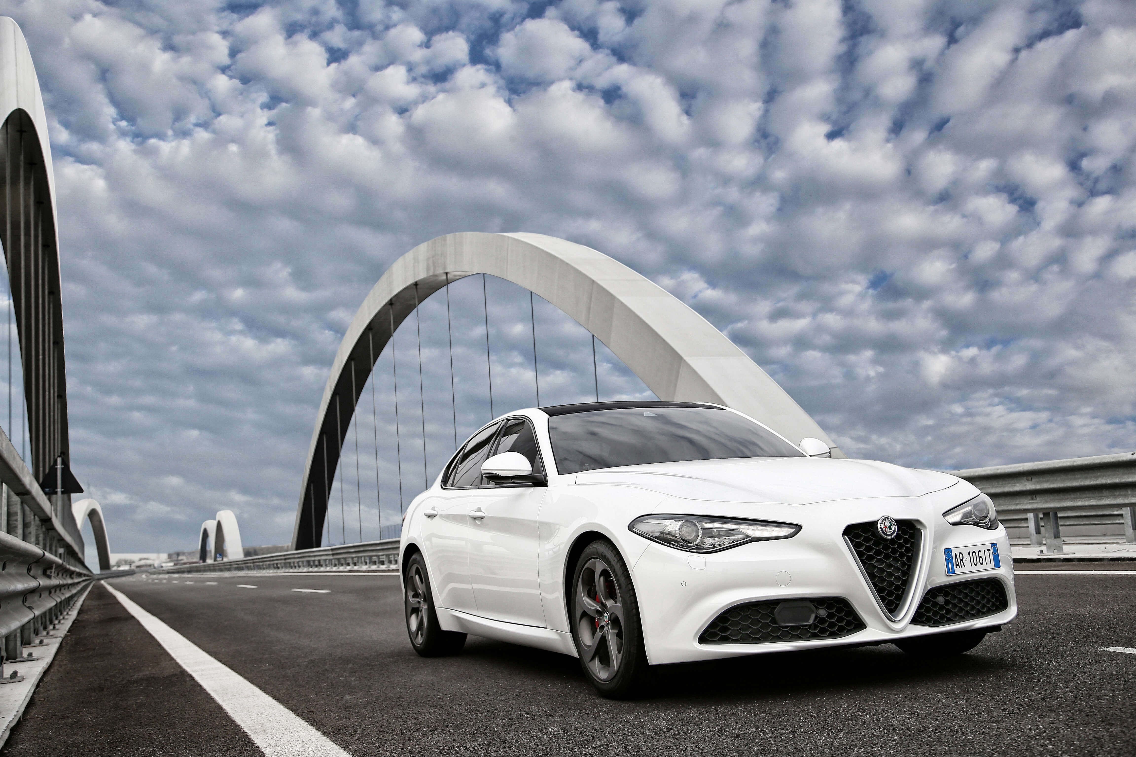 Alfa Romeo Giulia 桌布 壁紙 — 一架時尚的白色愛快羅密歐房車在戲劇性的多雲天空下自信地行駛在現代拱橋上。
