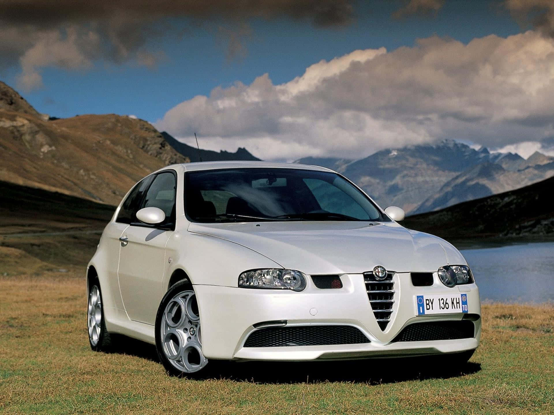 Alfa Romeo 147 桌布 壁紙 — 一架時尚的白色愛快羅密歐跑車停泊在草地上，背景是山脈和湖泊，天空多雲。