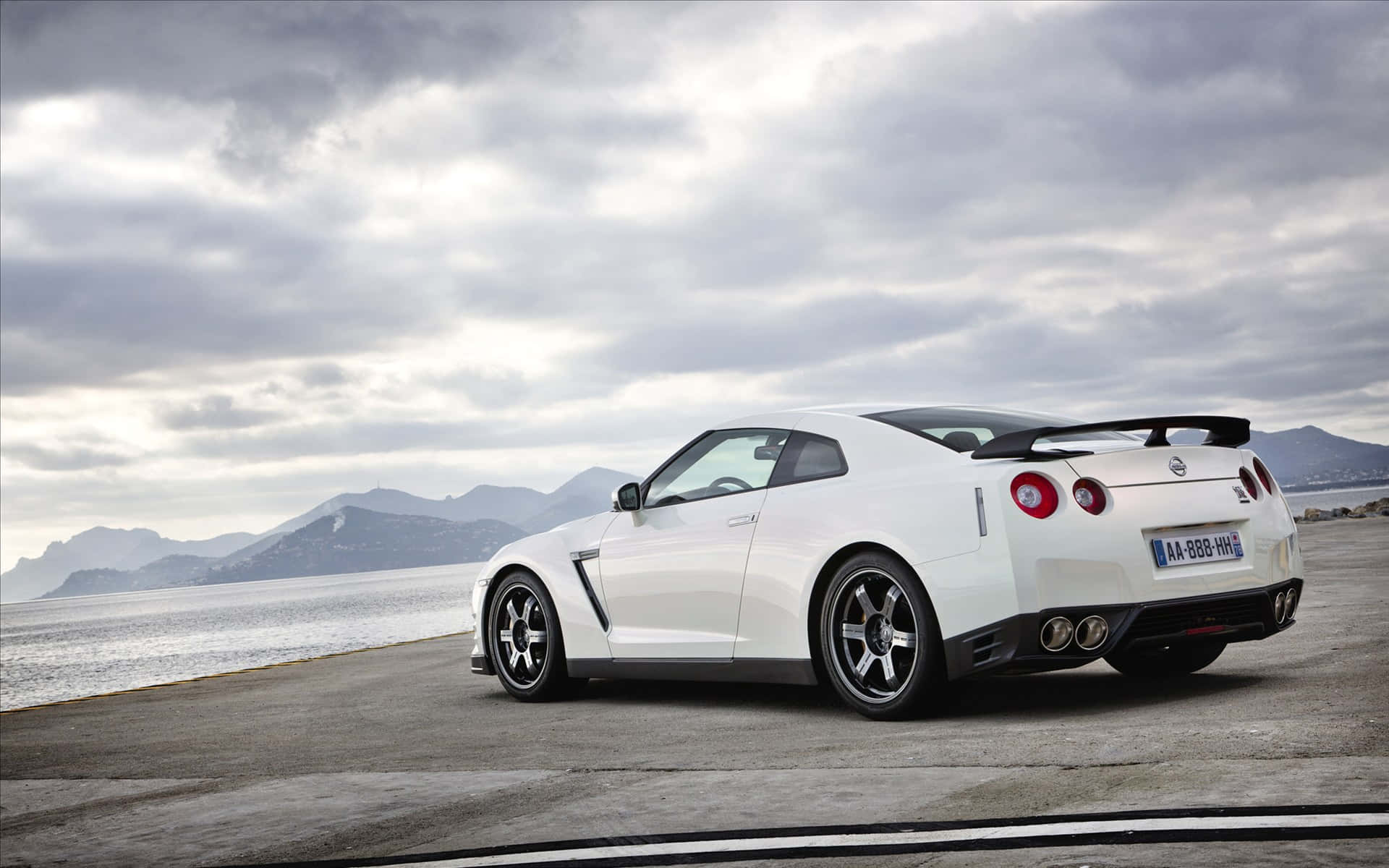 Gtr 桌布 壁紙 — 一架時尚的白色日產GT-R跑車停泊在沿海公路上，背景是山脈和陰天。