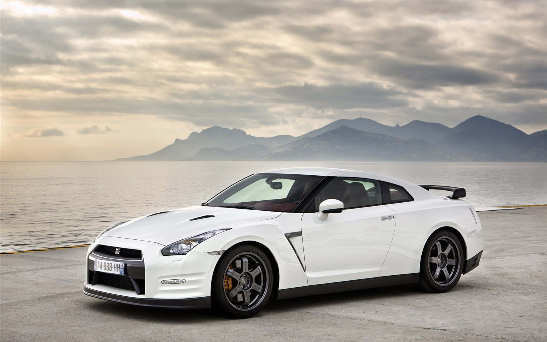 Gtr 桌布 壁紙 — 一架時尚的白色日產GT-R跑車停泊在沿海碼頭，背景有山脈和陰天。
