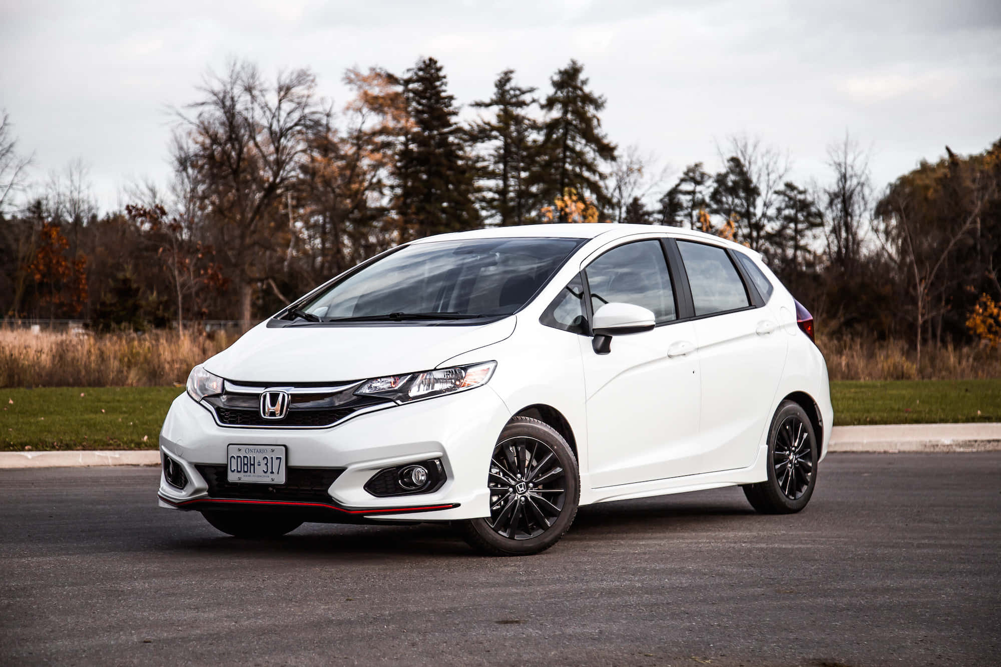 Honda Fit 桌布 壁紙 — 一架時尚的白色本田Fit，配黑色輪圈，停泊在瀝青路上，旁邊是秋天的樹木，天空多雲。