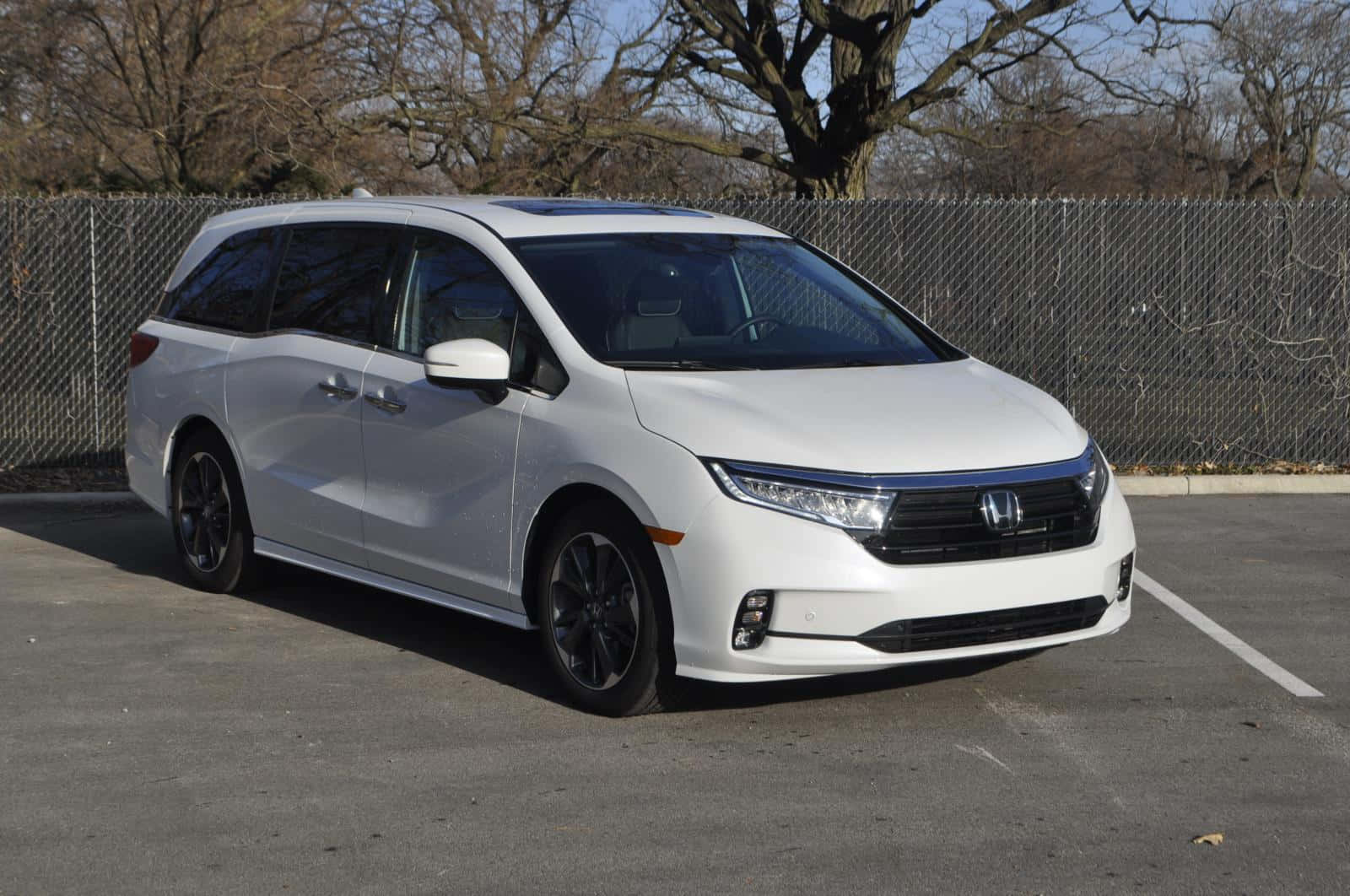 Honda Odyssey 桌布 壁紙 — 一架時尚的白色本田Odyssey小型貨車停泊在陽光明媚的瀝青停車場，背景有光禿禿的樹木和鐵絲網圍欄。