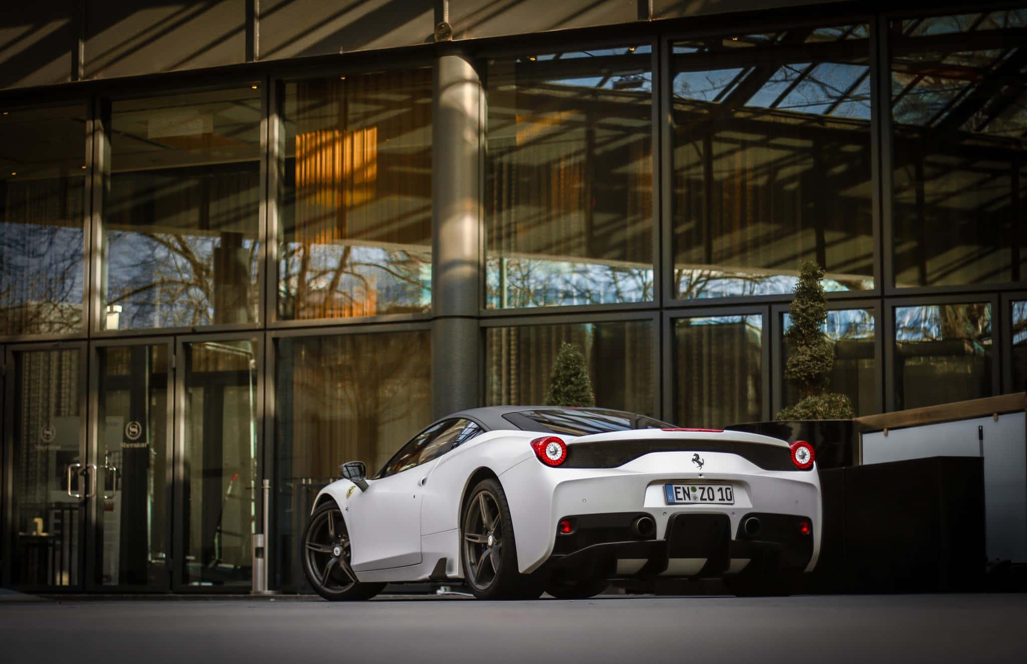 Ferrari 458 Speciale 桌布 壁紙 — 一架時尚的白色法拉利跑車停泊在現代玻璃建築外，陽光投射出溫暖的反射。
