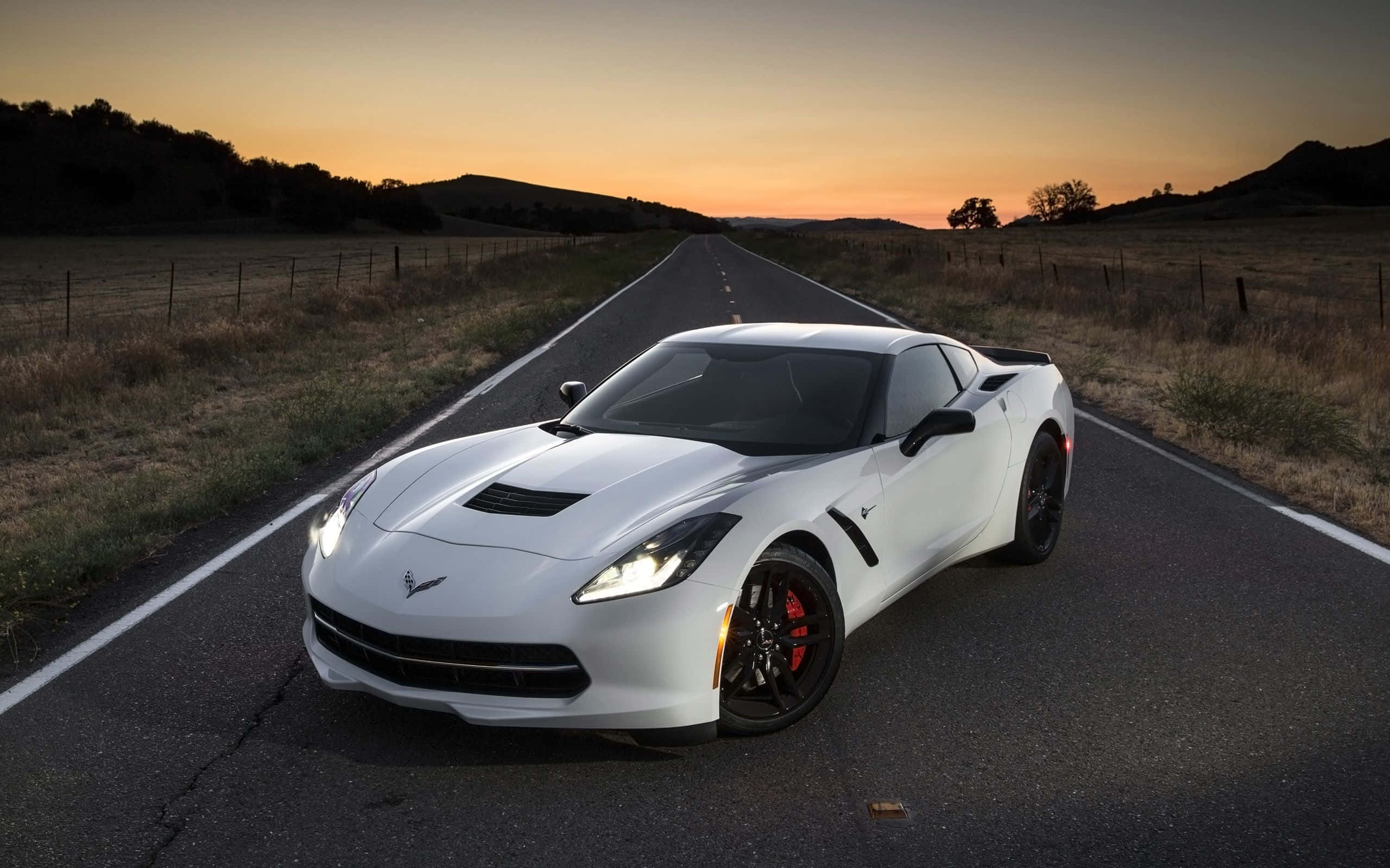 Chevrolet Corvette Stingray 桌布 壁紙 — 一架時尚的白色跑車，車頭燈閃爍，在日落時分停在一條漫長的鄉村道路上。