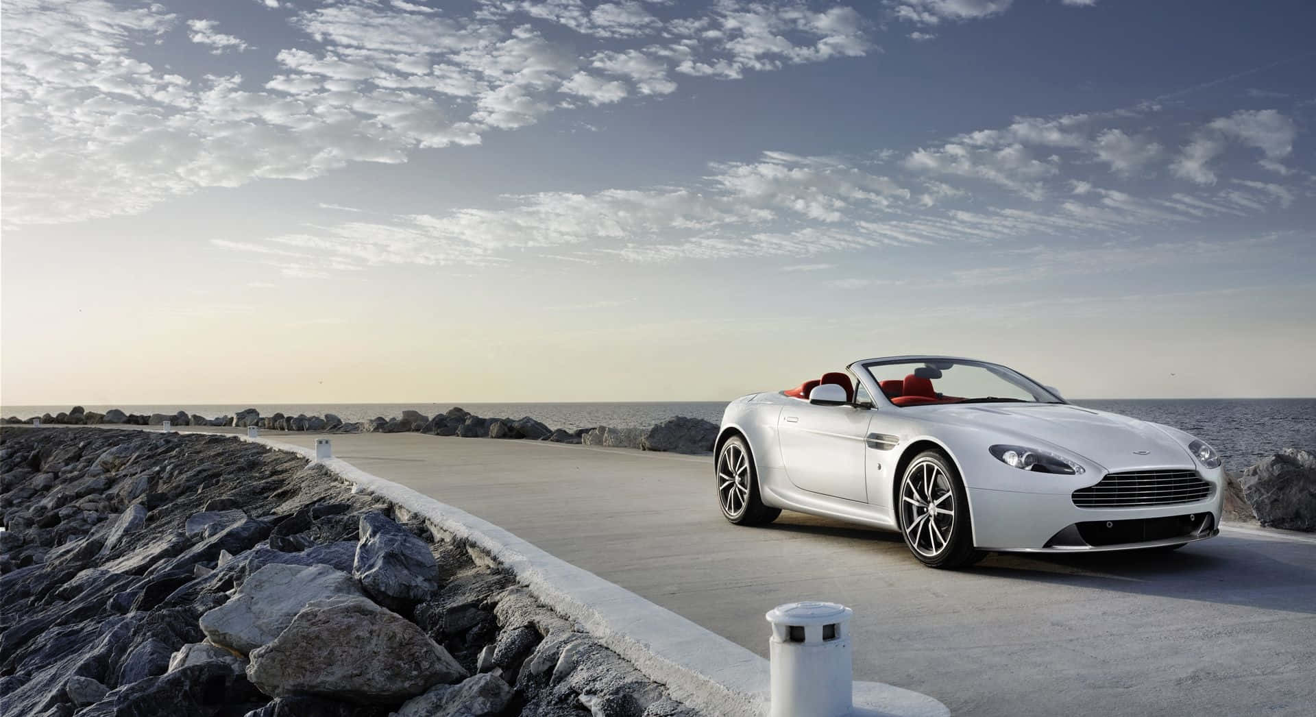 Aston Martin V12 Vantage 桌布 壁紙 — 一架時尚的白色開篷跑車，配備紅色內飾，沿著海岸道路行駛，旁邊是岩石峭壁和海洋，天空部分多雲。