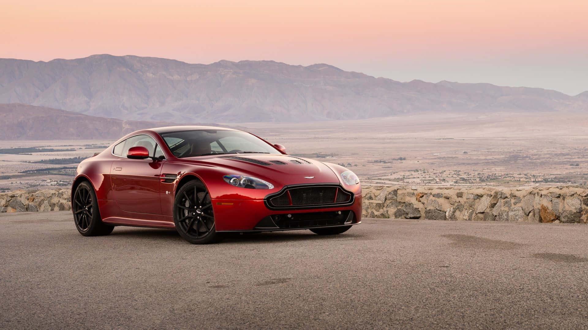 Aston Martin V12 Vantage 桌布 壁紙 — 一架時尚的紅色Aston Martin跑車停泊在鋪砌的觀景點上，背景是山脈和粉紅色的日落。