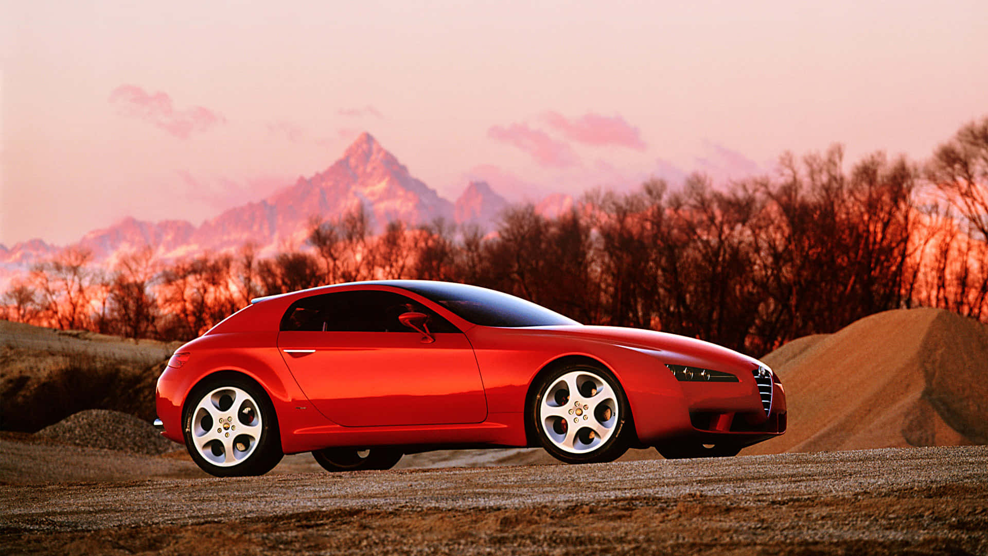 Alfa Romeo Brera 桌布 壁紙 — 一架時尚的紅色跑車停在碎石上，背景是白雪覆蓋的山脈和光禿禿的樹木在日落色調中閃耀。