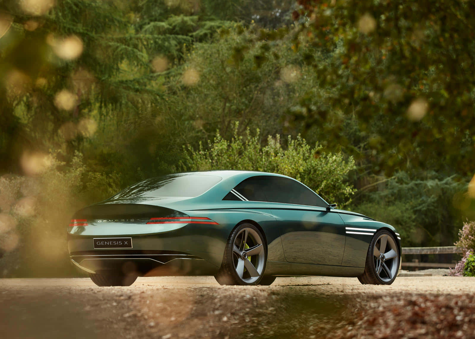 Genesis X Concept 桌布 壁紙 — 一架時尚的綠色 Genesis X 概念車停在碎石路上，前景有模糊的森林樹葉和金色散景。