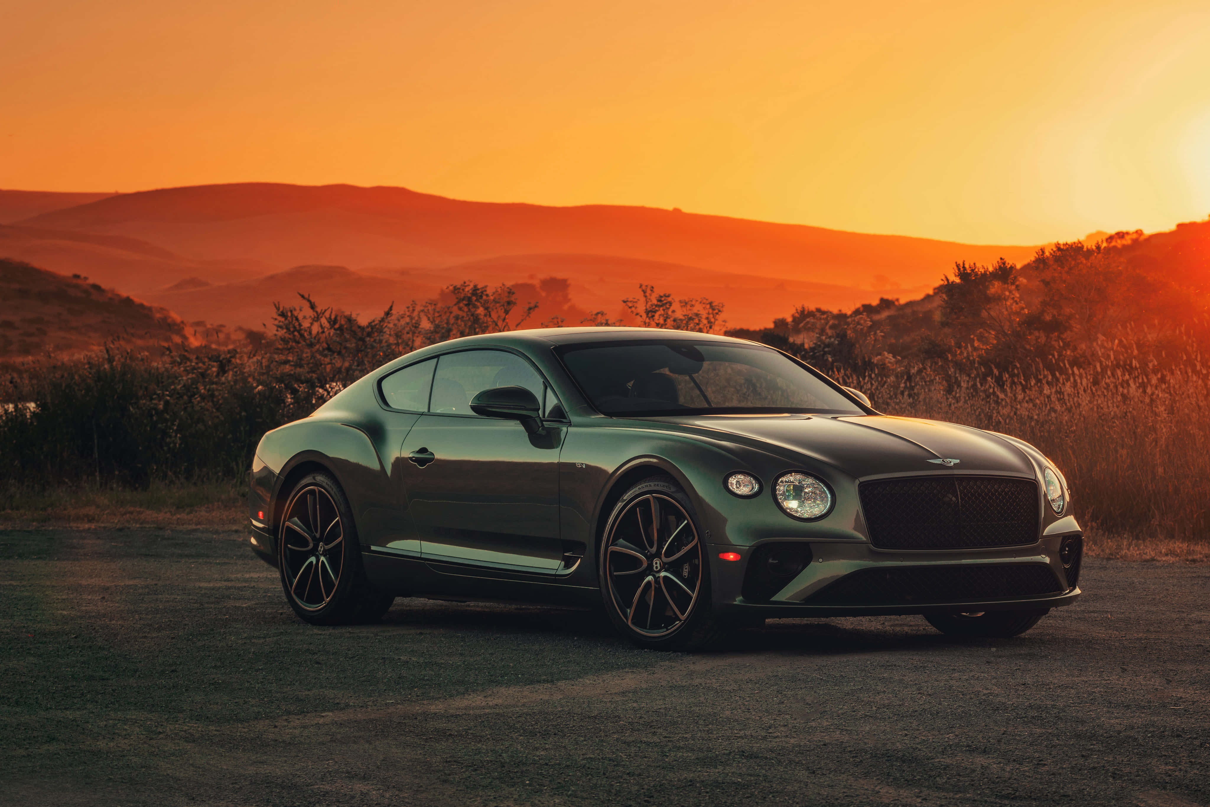 Bentley Continental Gt 桌布 壁紙 — 一架時尚的綠色賓利 Continental GT 停泊在日落時分的泥路上，起伏的山丘沐浴在溫暖的橙色光芒中。