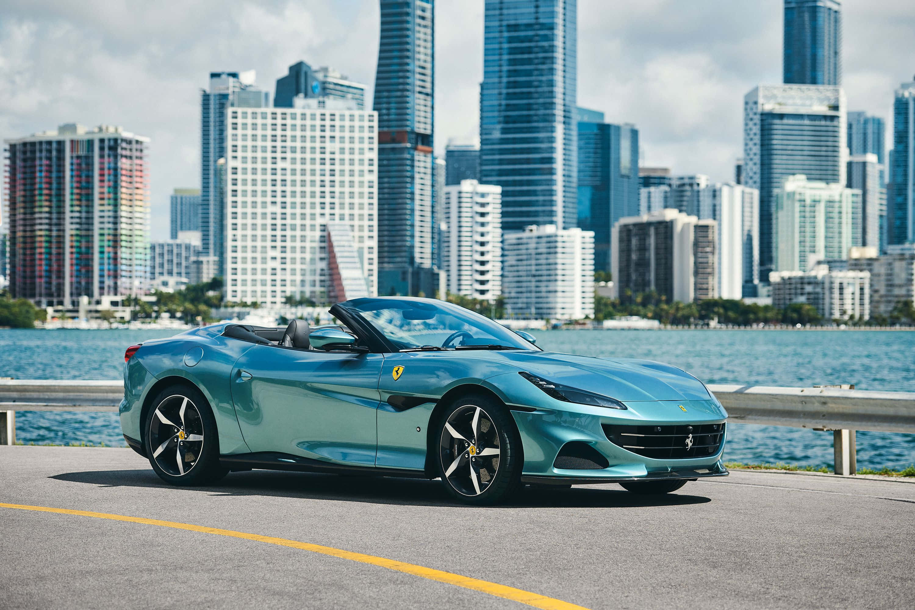 Ferrari Portofino 桌布 壁紙 — 一架時尚的藍綠色法拉利敞篷車停在海濱道路上，背後是現代城市天際線和藍色海水。