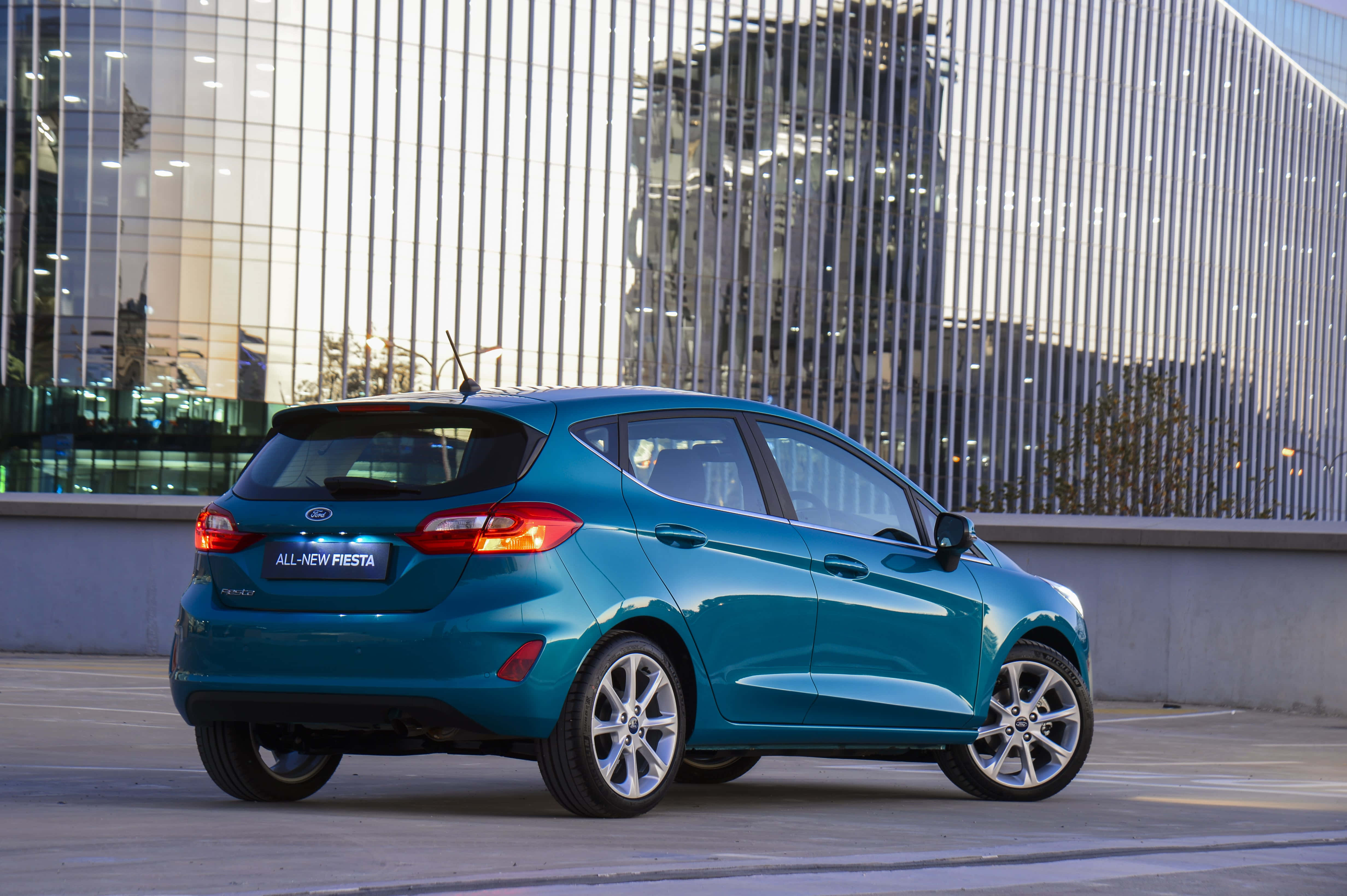 Compact Car 桌布 壁紙 — 一架時尚的藍綠色福特 Fiesta 停泊在現代玻璃建築前，建築帶有垂直金屬框架。