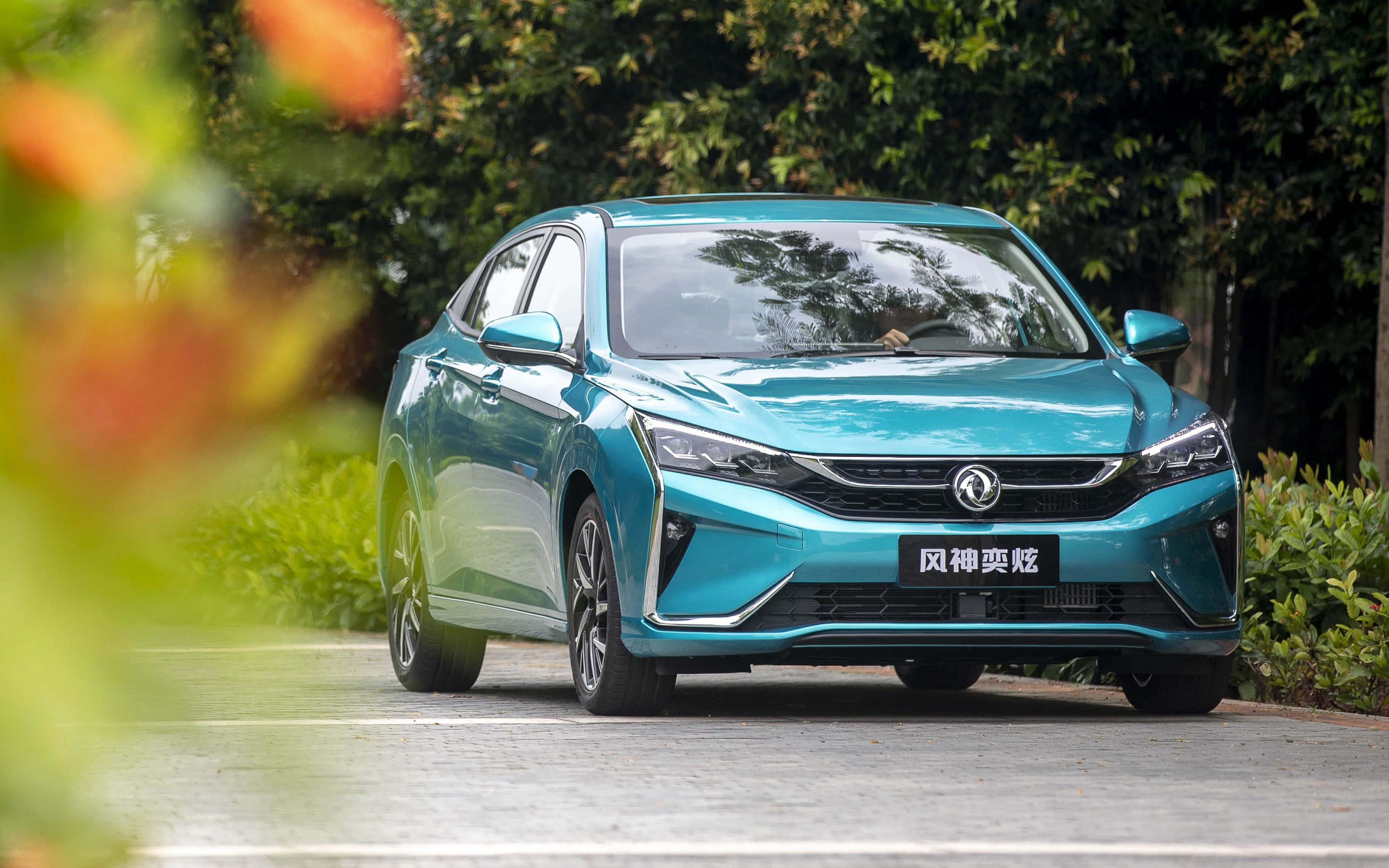 Dongfeng 桌布 壁紙 — 一架時尚的藍綠色轎車停泊在鋪砌的小路上，周圍環繞著茂密的綠色樹葉和模糊的橙色花朵。