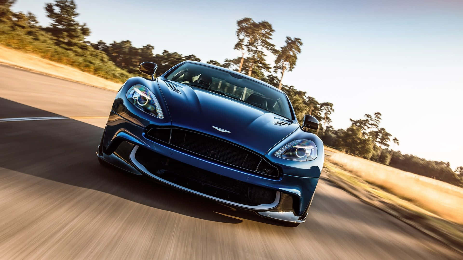 Aston Martin Vanquish 桌布 壁紙 — 一架時尚的藍色 Aston Martin 跑車在陽光明媚的鄉村道路上疾馳，背景有模糊的動感和樹木。