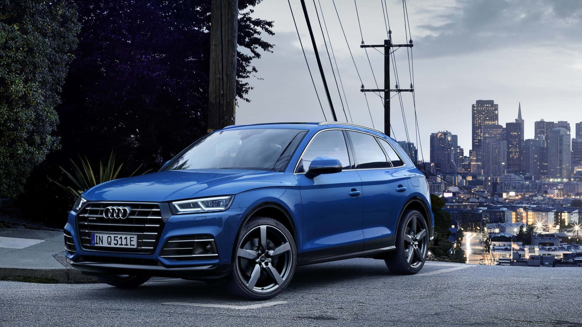 Audi Q5 桌布 壁紙 — 一架時尚的藍色 Audi Q5 SUV 停泊在城市的山坡上，俯瞰著暮色中燈火通明的天際線。