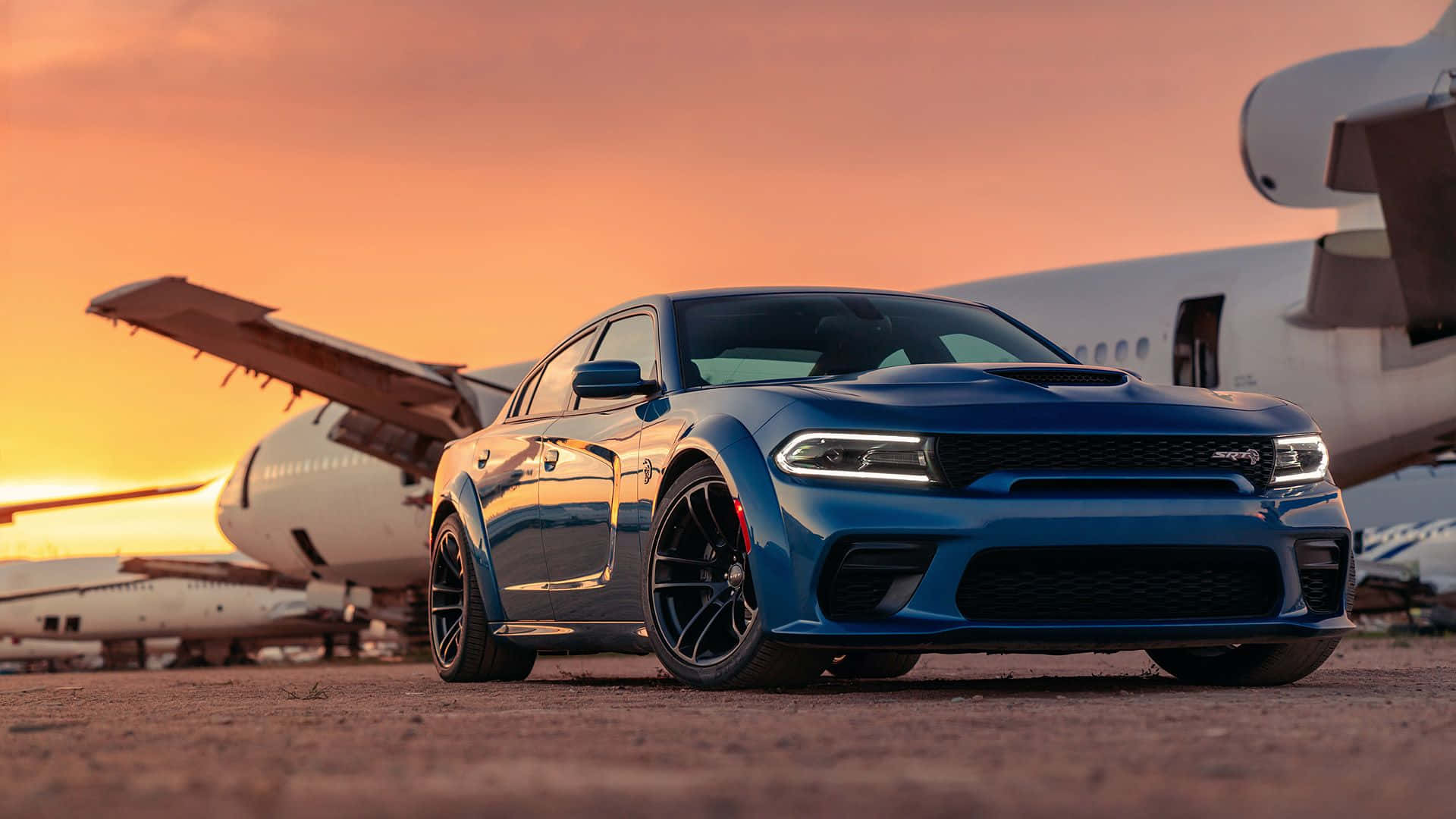 Dodge Charger 桌布 壁紙 — 一架時尚的藍色 Dodge Charger Hellcat 停泊在停機坪上，背景是大型飛機，正值熾熱的日落時分。