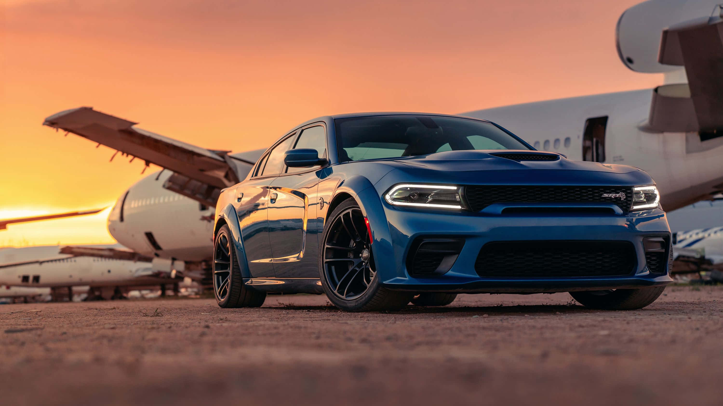 Dodge Charger 桌布 壁紙 — 一架時尚的藍色 Dodge Charger SRT Hellcat 停泊在停機坪上，背景是大型噴射機，正值熾熱的日落時分。