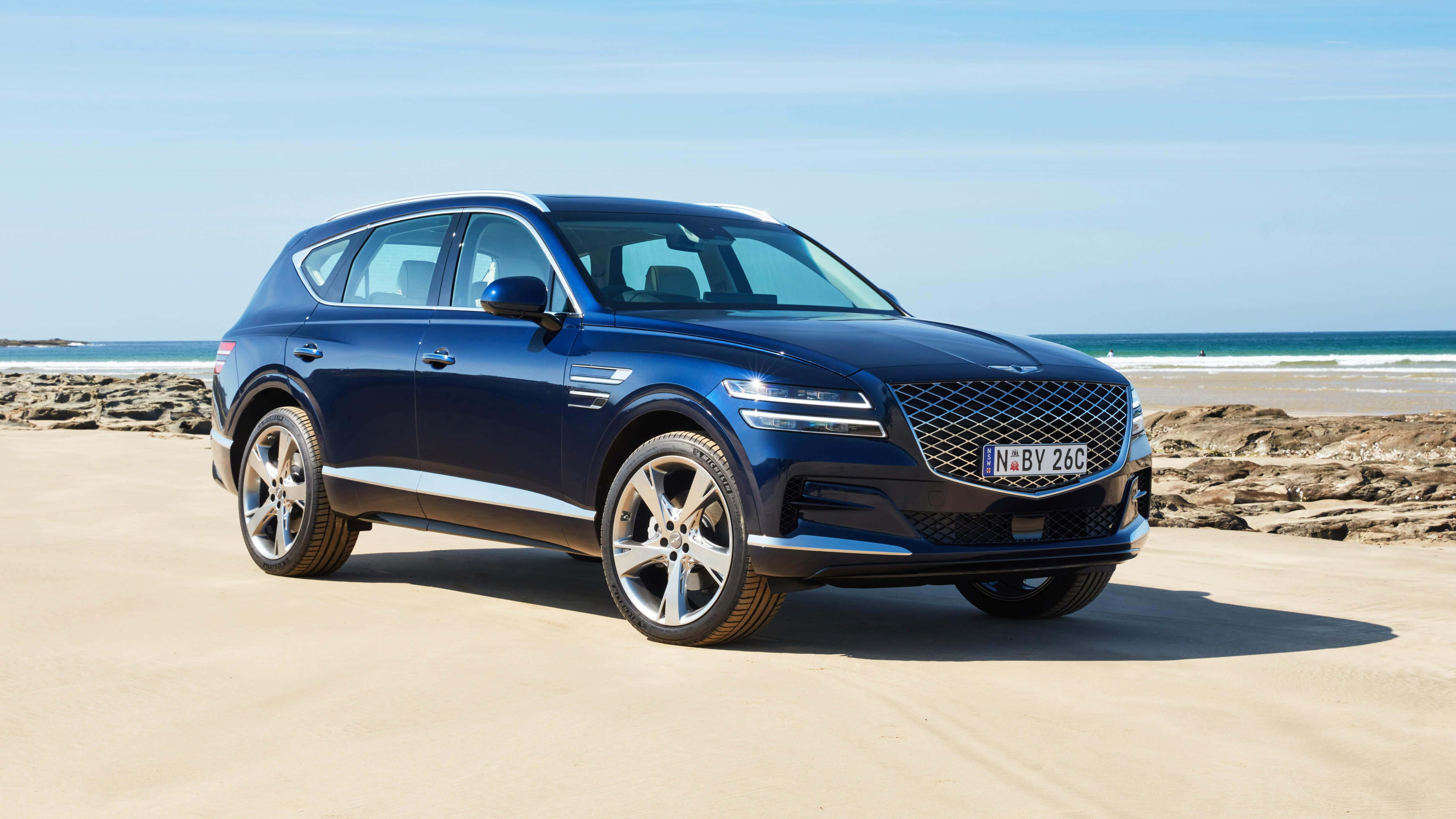 Genesis Essentia 桌布 壁紙 — 一架時尚的藍色 Genesis GV80 SUV 停泊在陽光明媚的沙灘上，背景是海浪和岩石海岸。