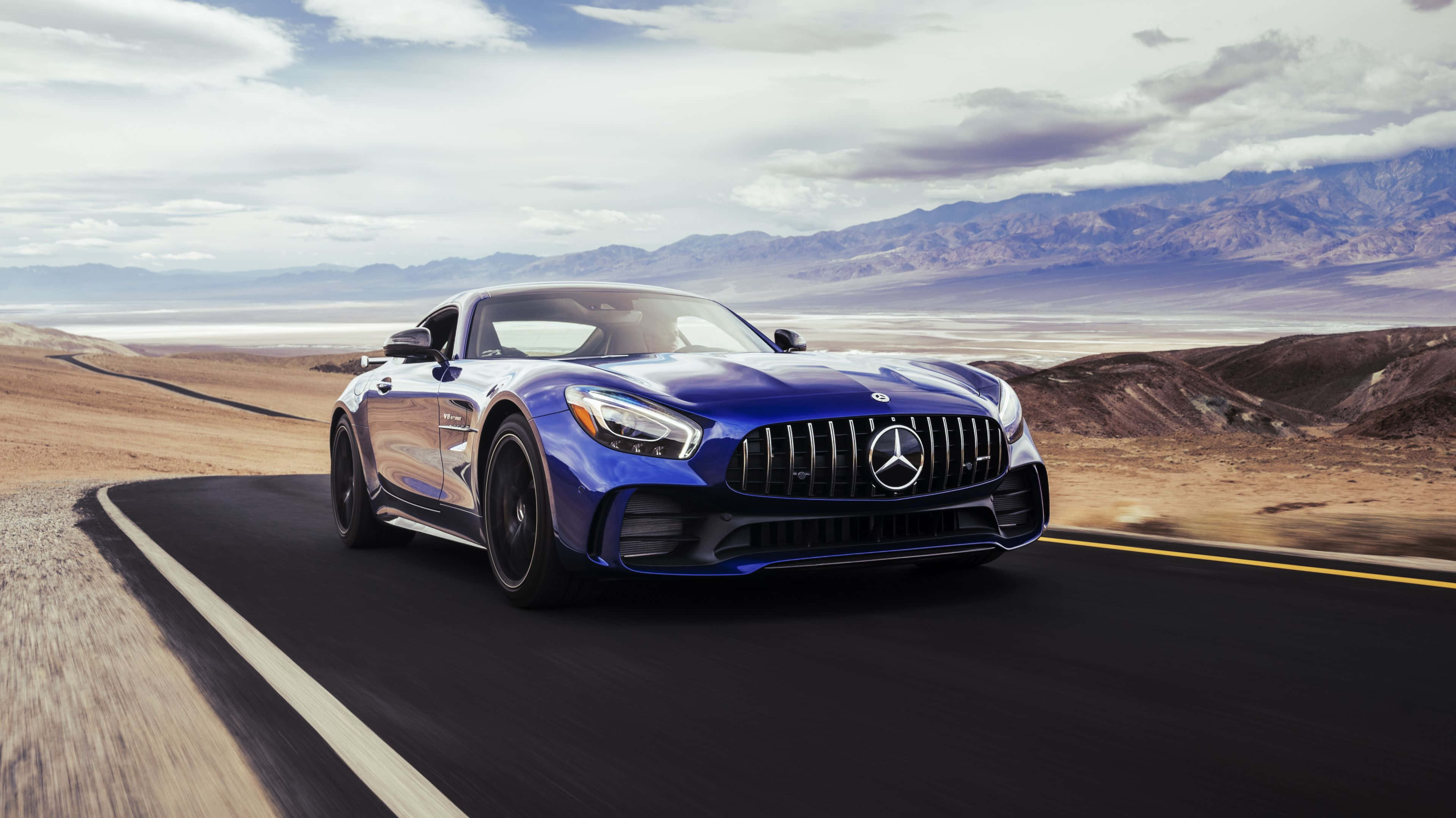 4K Mercedes 桌布 壁紙 — 一架時尚的藍色 Mercedes-AMG GT 跑車在沙漠公路上疾馳，遠方山巒起伏，天空多雲。