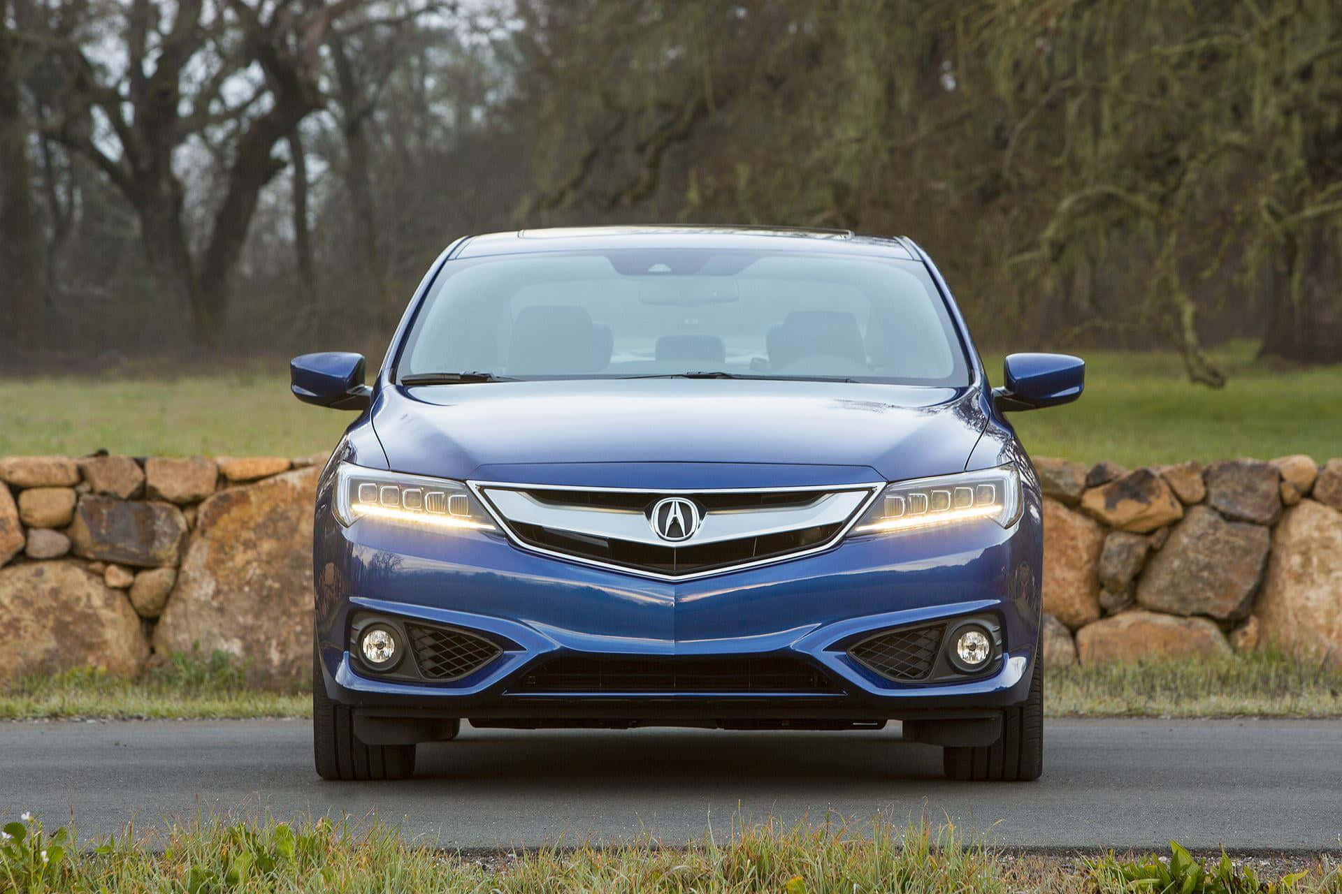Acura Ilx 桌布 壁紙 — 一架時尚的藍色Acura房車在由石牆和光禿樹木環繞的瀝青路上向前行駛。