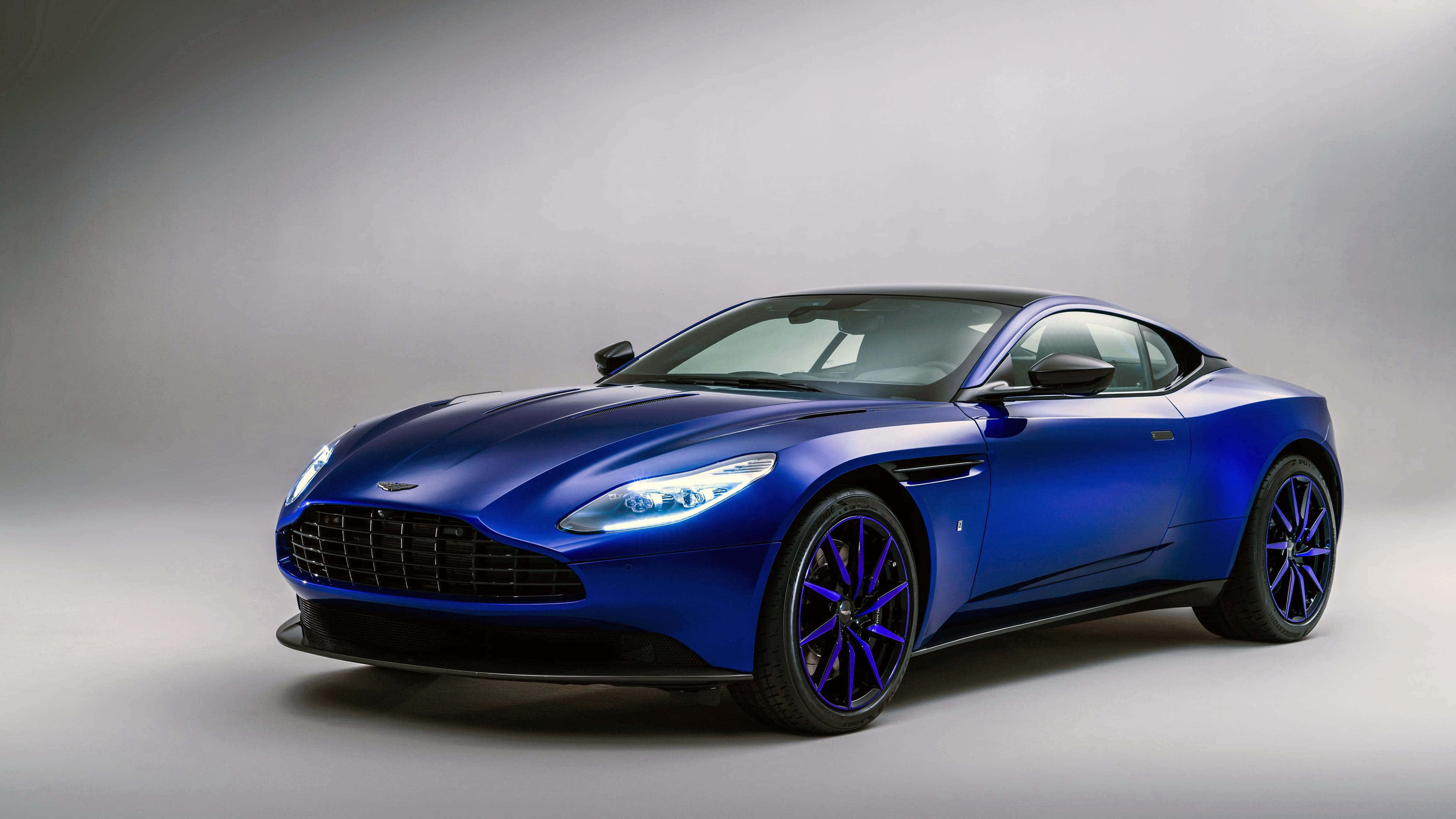 Aston Martin Db11 桌布 壁紙 — 一架時尚的藍色Aston Martin跑車，配備發光的頭燈和紫色輪圈，在中性工作室背景前擺放。