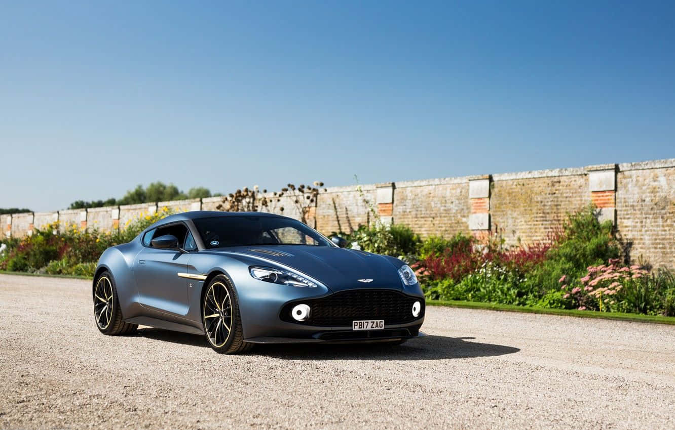 Aston Martin Vanquish 桌布 壁紙 — 一架時尚的藍色Aston Martin跑車停泊在碎石車道上，旁邊是磚牆和花園，在晴朗的藍天下盛開著花朵。
