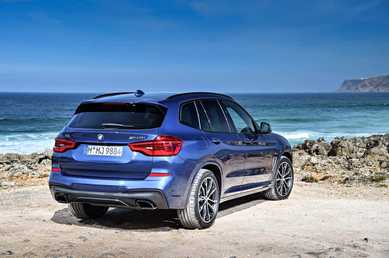Bmw X3 桌布 壁紙 — 一架時尚的藍色BMW X3 M40i SUV停泊在岩石海岸懸崖上，俯瞰海洋，天空湛藍清澈。