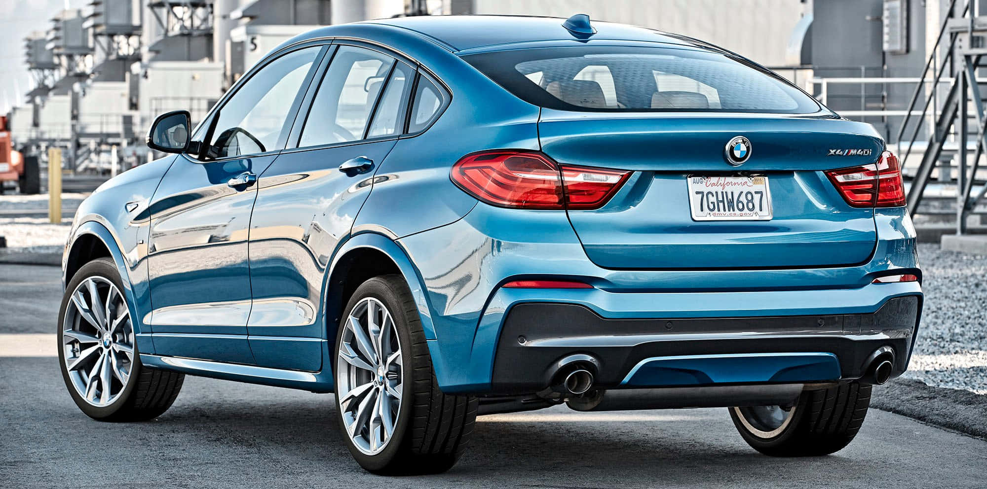 Bmw X4 桌布 壁紙 — 一架時尚的藍色BMW X4 M40i SUV停泊在瀝青路上，背景是工業建築物。