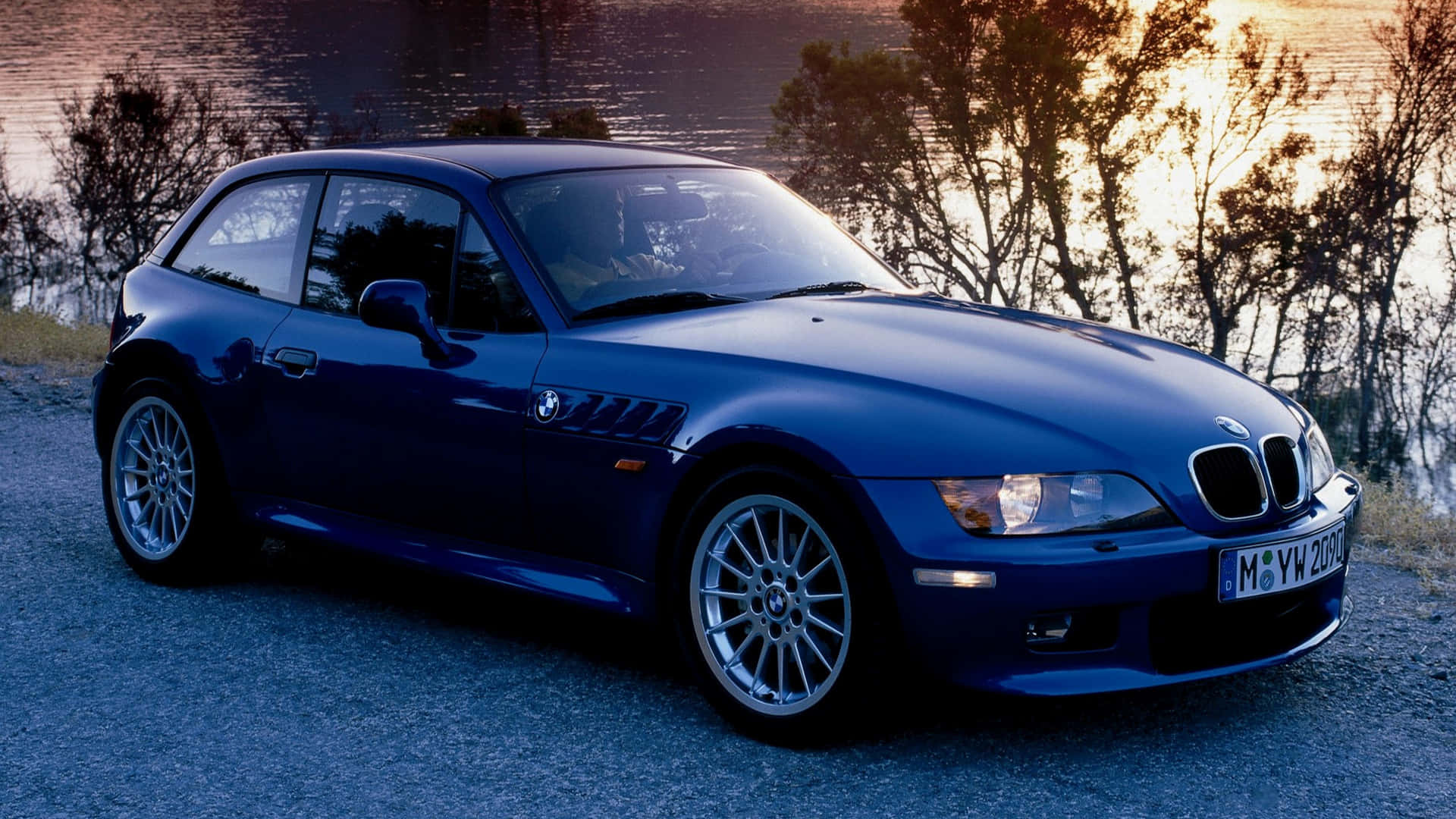 Bmw Z3 桌布 壁紙 — 一架時尚的藍色BMW Z3跑車停泊在寧靜的湖邊，金色夕陽下樹木呈現剪影。