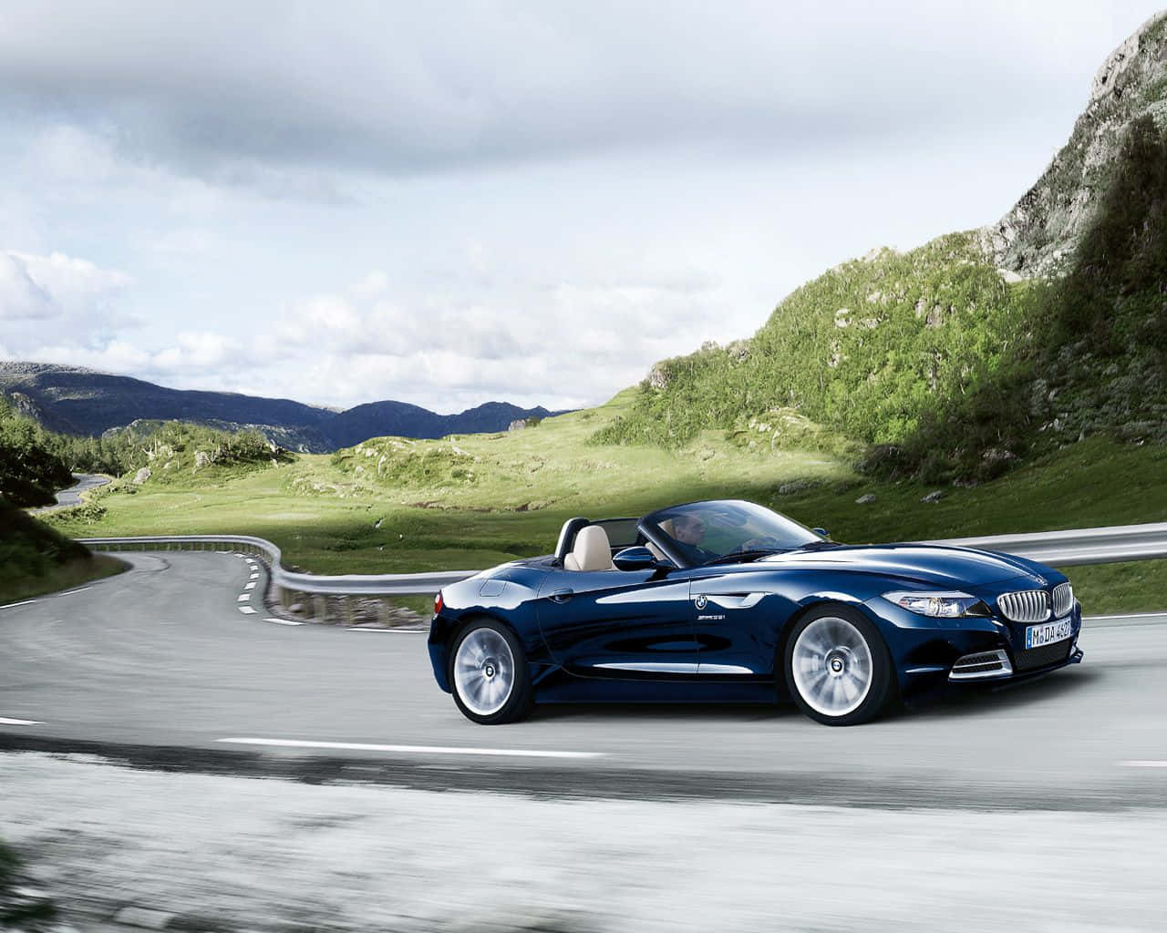 Bmw Z4 桌布 壁紙 — 一架時尚的藍色BMW Z4開篷跑車在陰天之下，沿著蜿蜒的山路穿過翠綠的山丘飛馳。