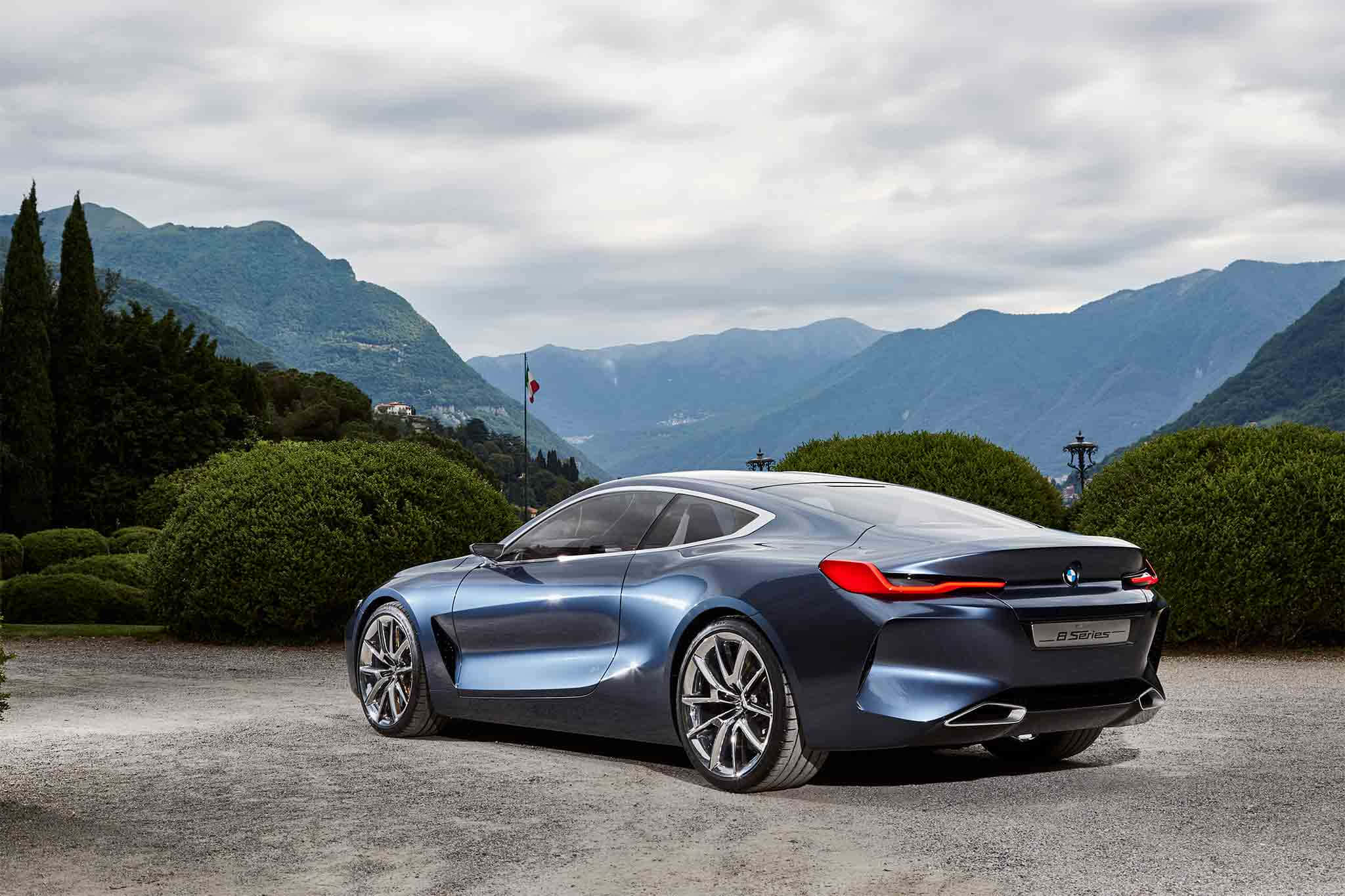 Bmw 8 Series 桌布 壁紙 — 一架時尚的藍色BMW概念車停在碎石上，背景有山脈和修剪整齊的樹籬，天空多雲。