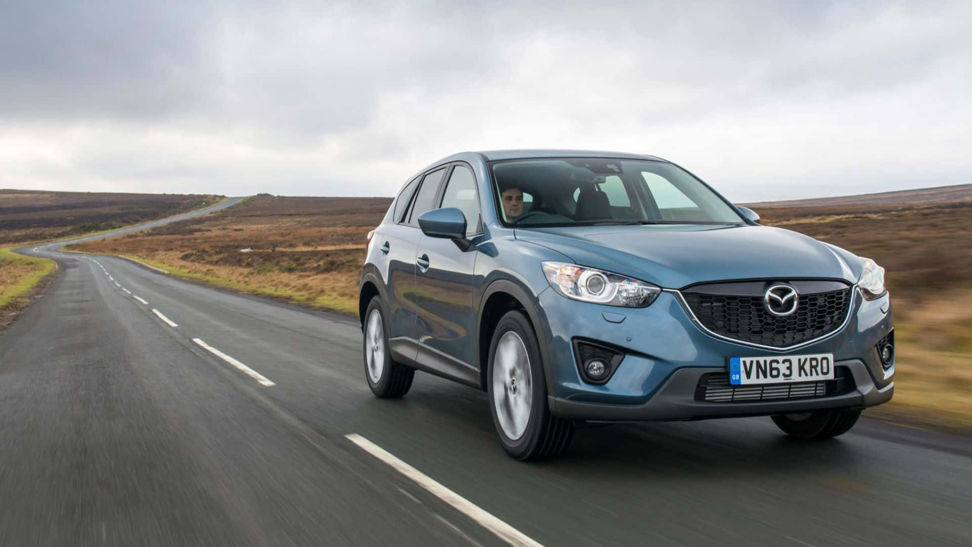 Mazda Cx-5 桌布 壁紙 — 一架時尚的藍色Mazda CX-5在陰天下行駛於蜿蜒的鄉村道路，穿過開闊的荒野。