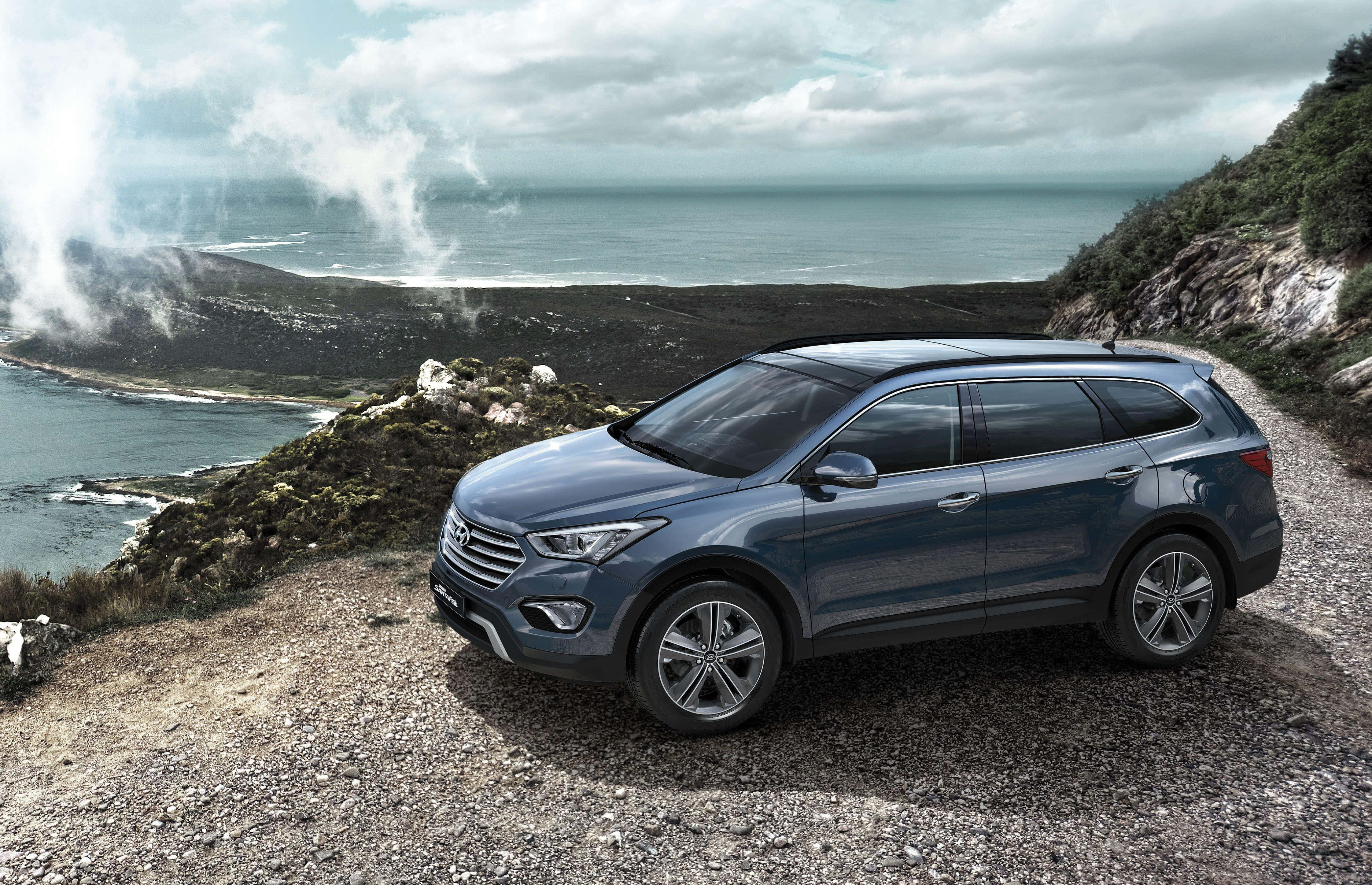 Hyundai Santa Fe 桌布 壁紙 — 一架時尚的藍色SUV停泊在崎嶇的海岸懸崖上，俯瞰著霧氣瀰漫的海洋，上方有戲劇性的雲層。