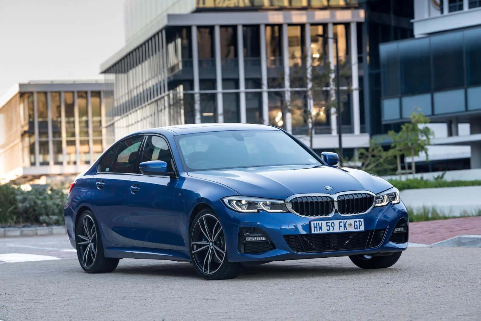 Bmw 3 Series 桌布 壁紙 — 一架時尚的藍色寶馬房車停泊在現代玻璃建築前，陽光在其拋光表面反射。