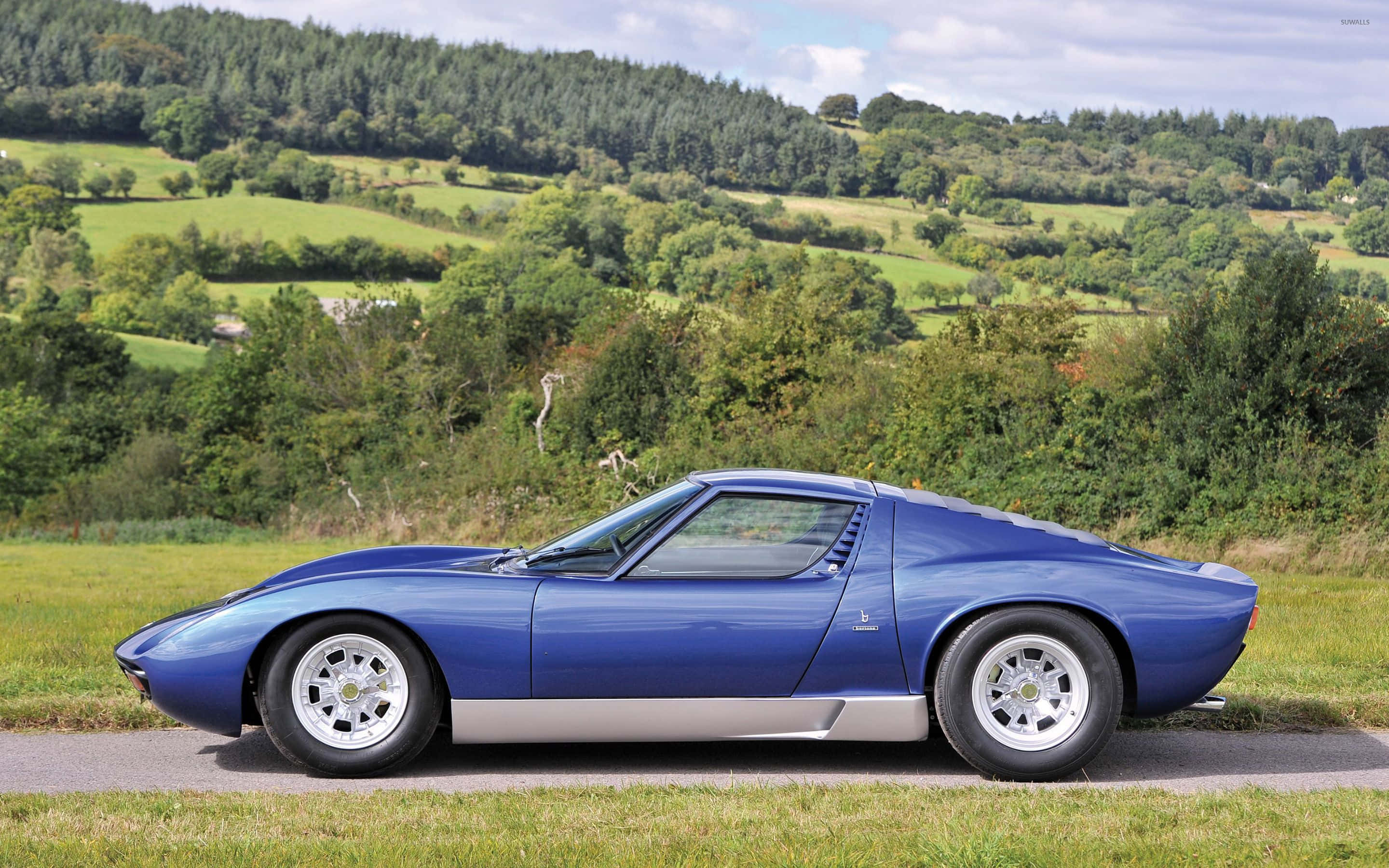 Lamborghini Miura 桌布 壁紙 — 一架時尚的藍色林寶堅尼Miura跑車停泊在鄉村道路上，背景是起伏的綠色山丘和茂密的森林，天空部分多雲。