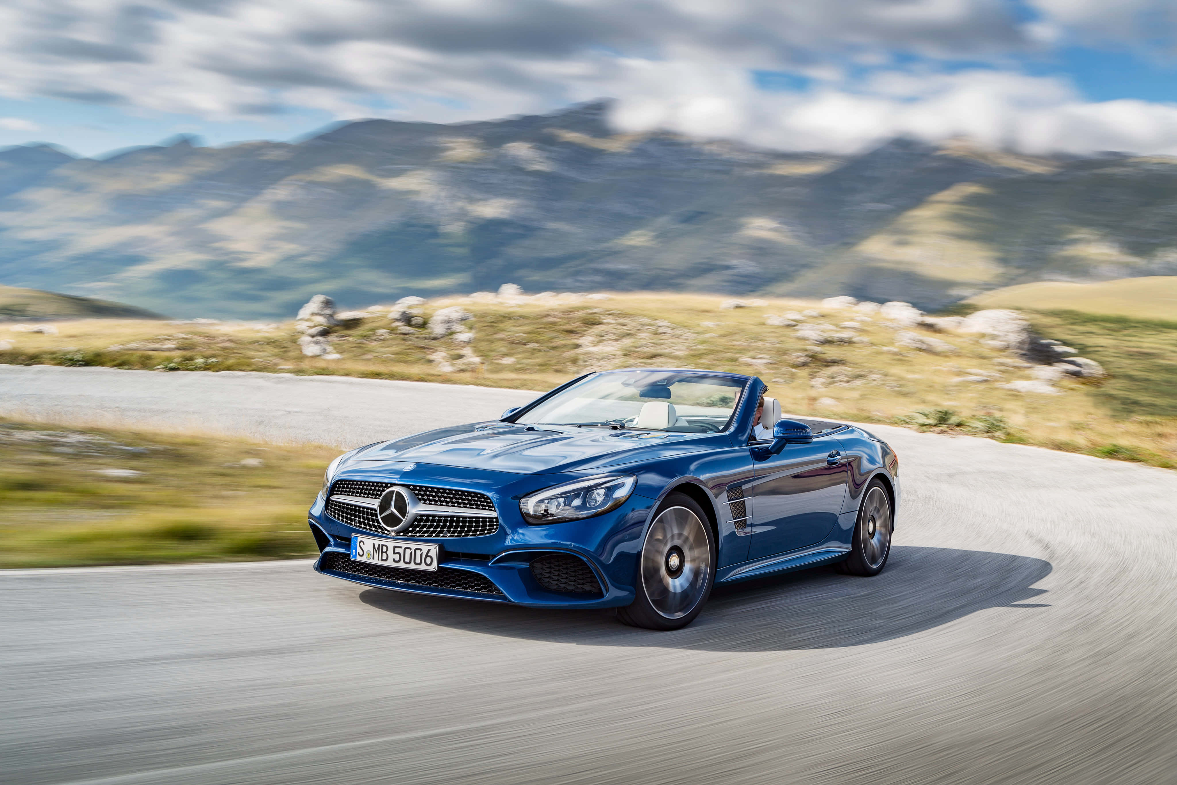 Mercedes Benz Sl-class 桌布 壁紙 — 一架時尚的藍色梅賽德斯奔馳敞篷車在部分多雲的天空下行駛在蜿蜒的山路上。