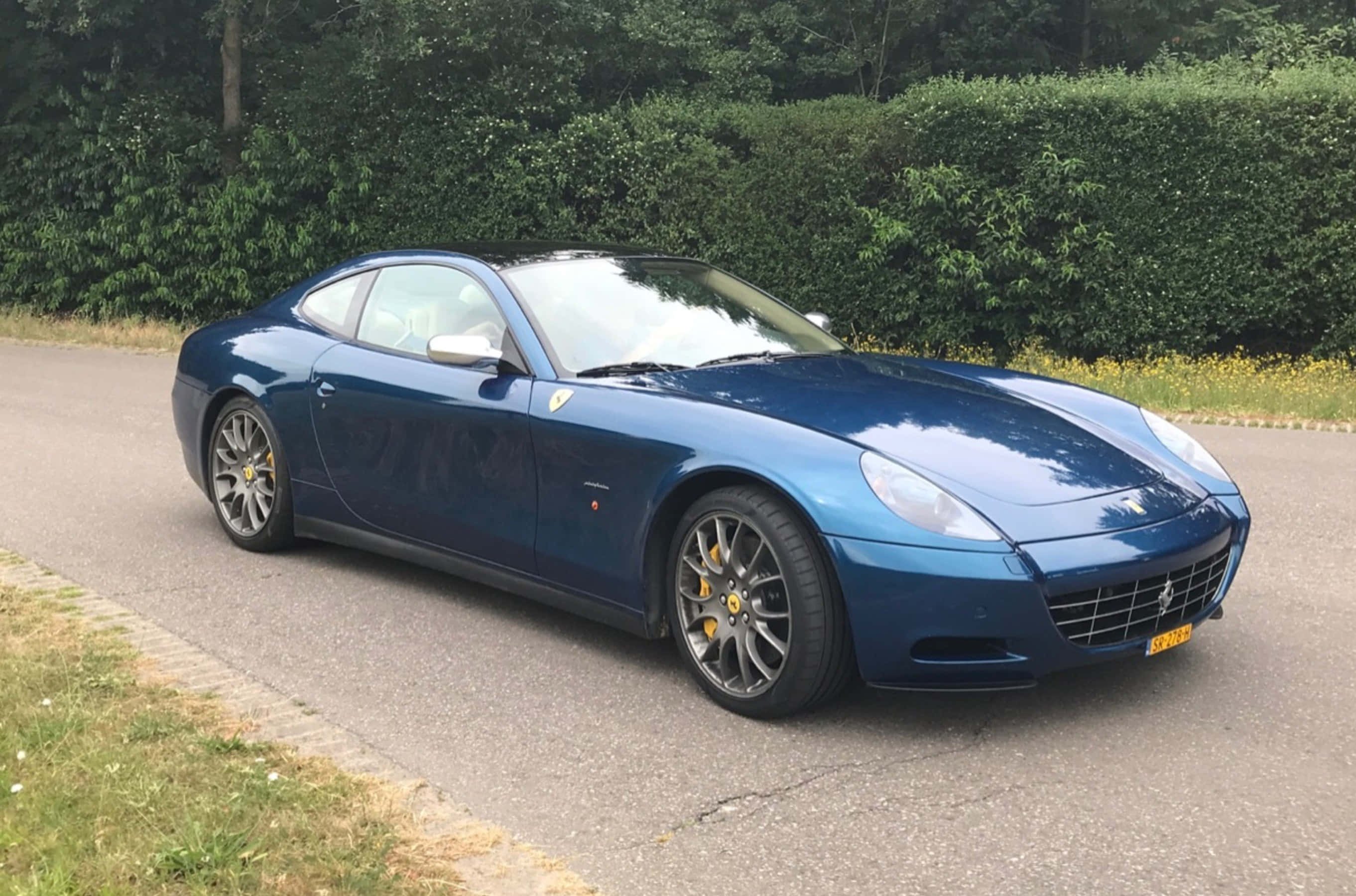 Ferrari 612 Scaglietti 桌布 壁紙 — 一架時尚的藍色法拉利612 Superamerica停泊在鋪砌的道路上，旁邊有綠色籬笆和草地。