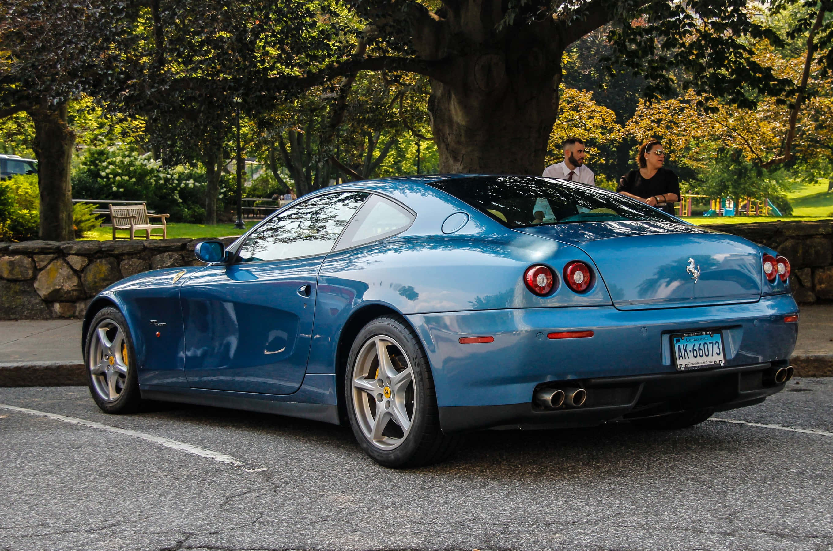 Ferrari 612 Scaglietti 桌布 壁紙 — 一架時尚的藍色法拉利停泊在樹木林立的公園裡，附近有幾個人坐在陽光普照的日子裡。