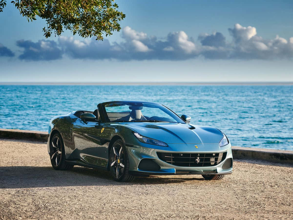 Ferrari Portofino 桌布 壁紙 — 一架時尚的藍色法拉利開篷車停泊在陽光明媚的海灘上，背景是碧綠的海洋和散落的雲朵。