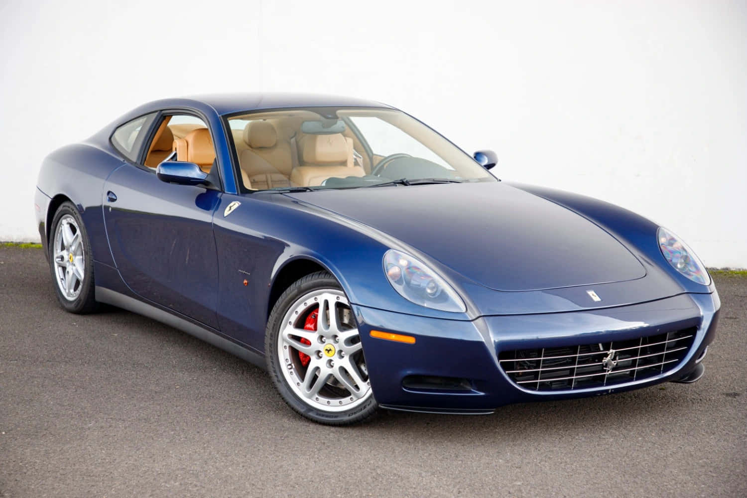 Ferrari 612 Scaglietti 桌布 壁紙 — 一架時尚的藍色法拉利雙門跑車，配備淺褐色皮革內飾，停泊在瀝青路面上，背景是純白色牆壁。